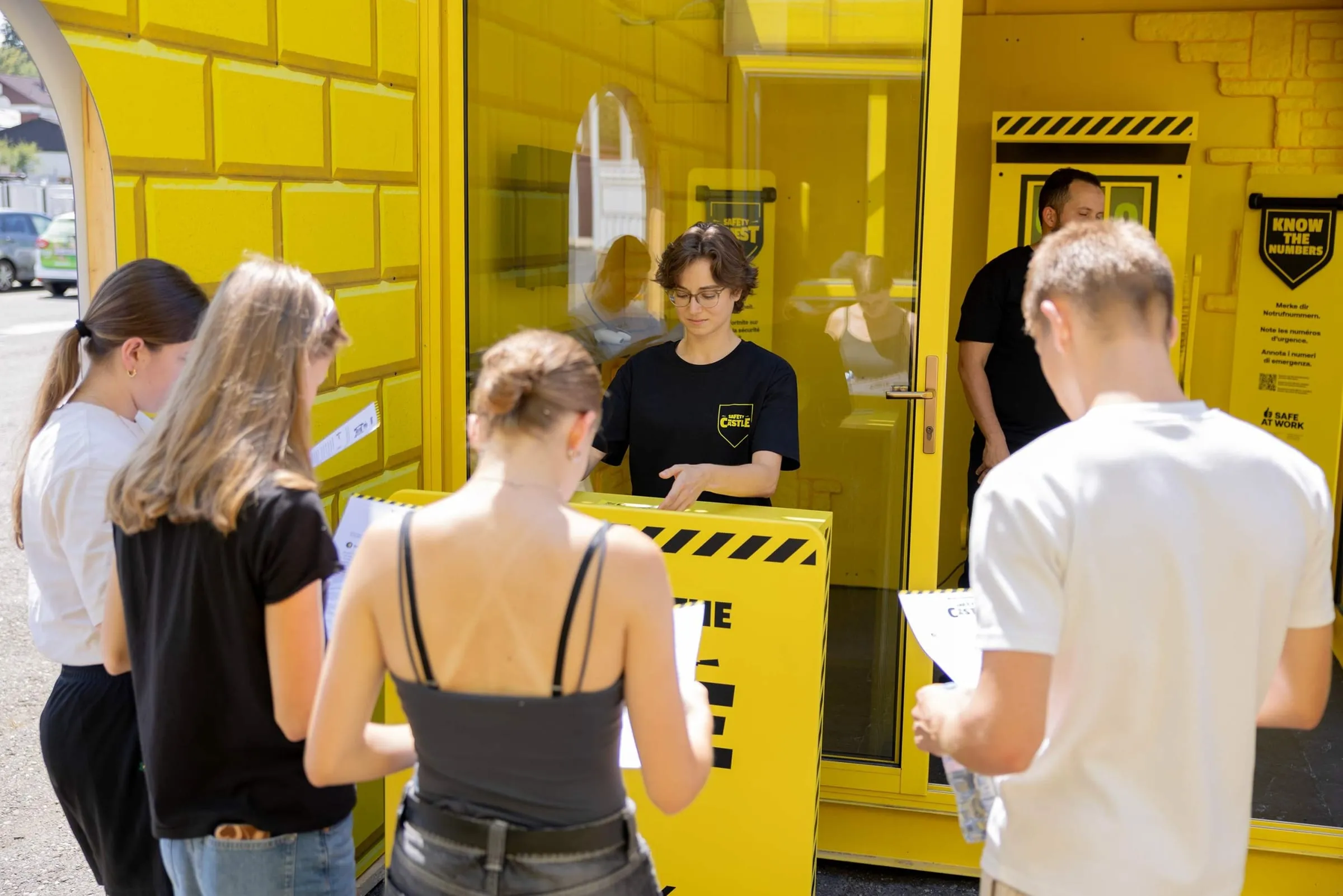 Four apprentices with registration forms at the yellow desk, a promoter in a Safety Castle shirt in front of the container behind them