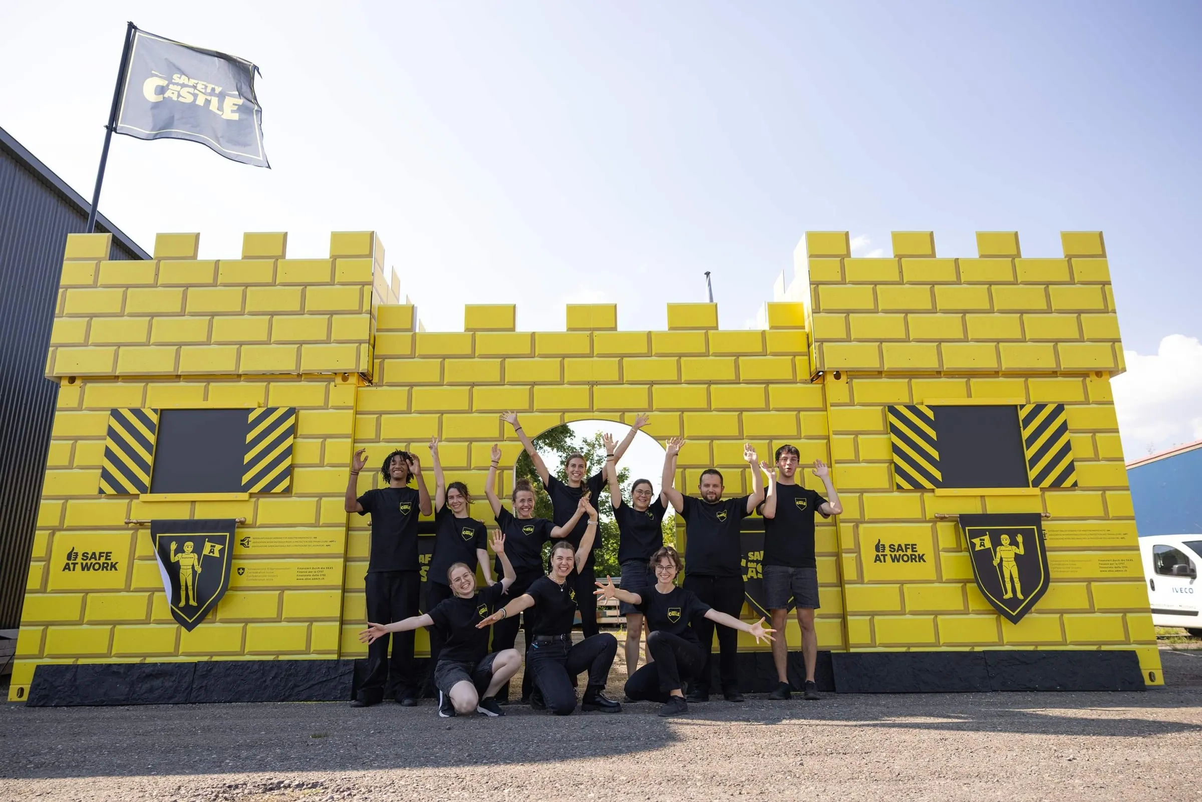 Team in black T-shirts cheering in front of the yellow Safety Castle with battlements, a knight crest and the Safe at Work logo, the flag above