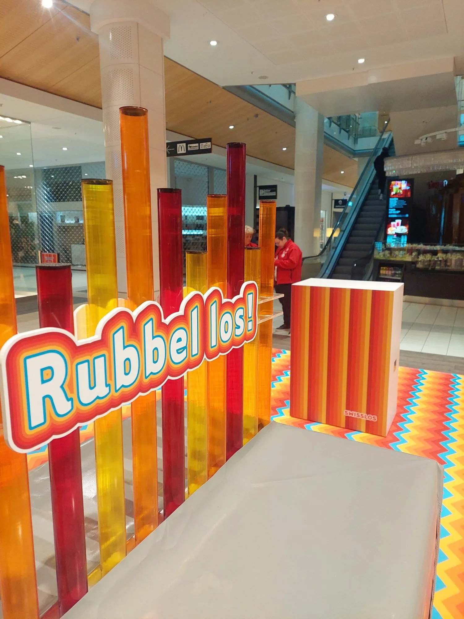 Schild «Rubbel los!» vor roten, orangen und gelben Acrylstäben, dahinter gestreifter Swisslos-Counter