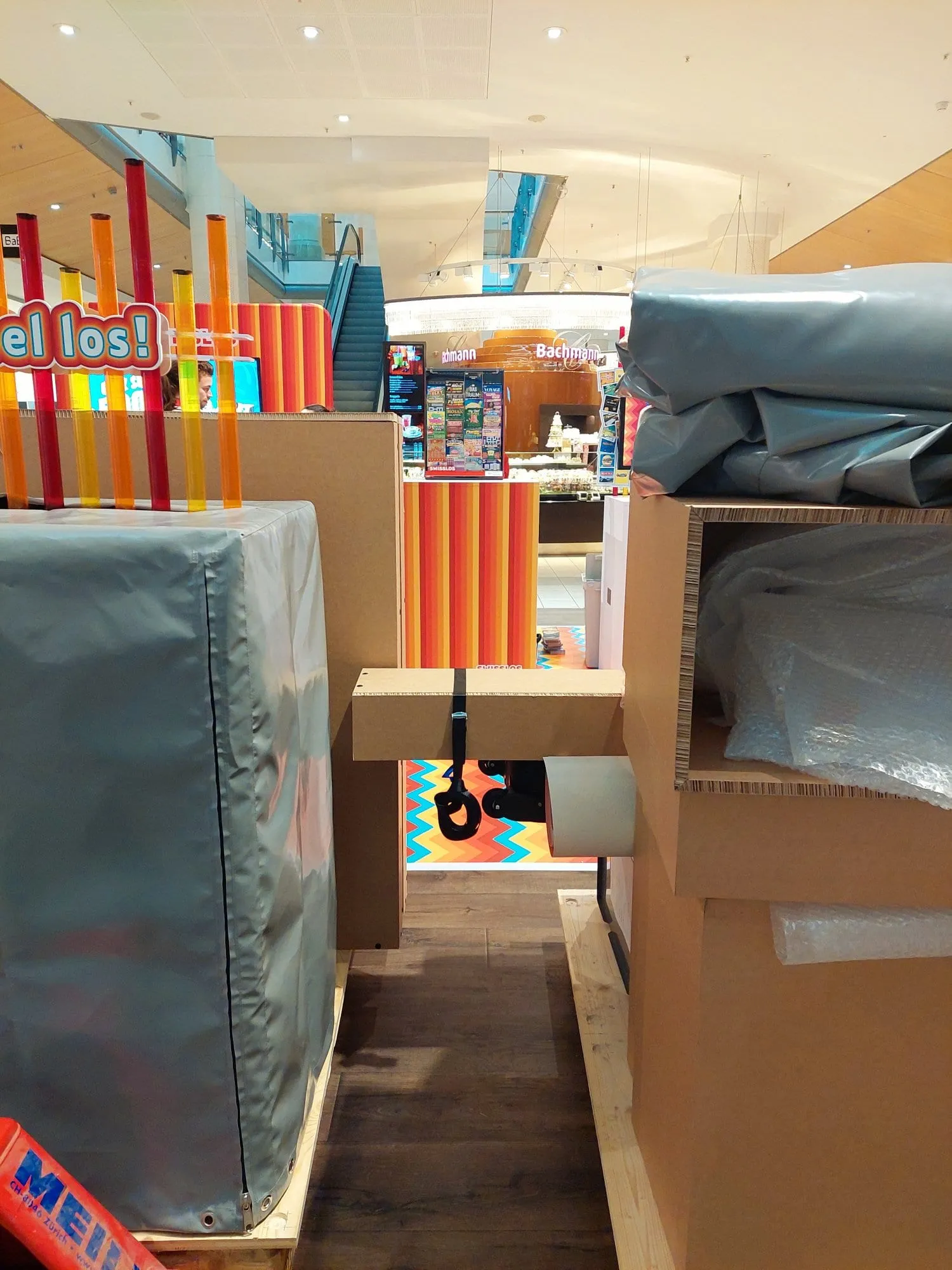 Aufbau des Swisslos-Stands: verpackte Elemente mit grauer Plane, Rubbel-los-Schild und bunte Acrylstäbe im Einkaufszentrum