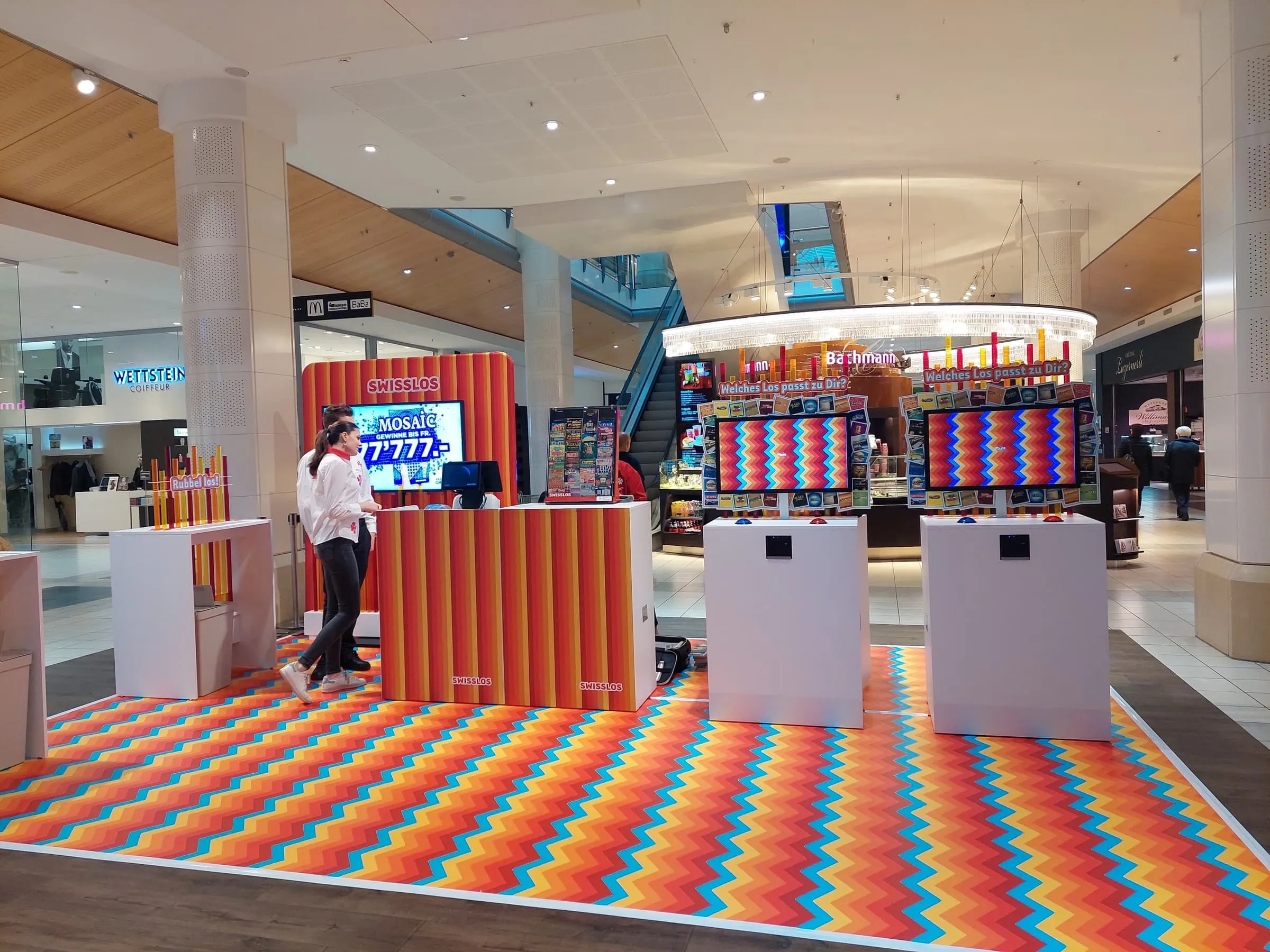 Swisslos stand with a zigzag carpet, orange striped counter and two screen-based game stations in a shopping centre