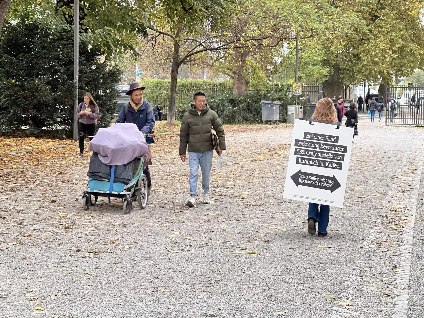 Sandwichfrau mit Oatly-Schild «Gratis Kaffee mit Oatly» auf einem Kiesweg im Park, Passanten mit Kinderwagen