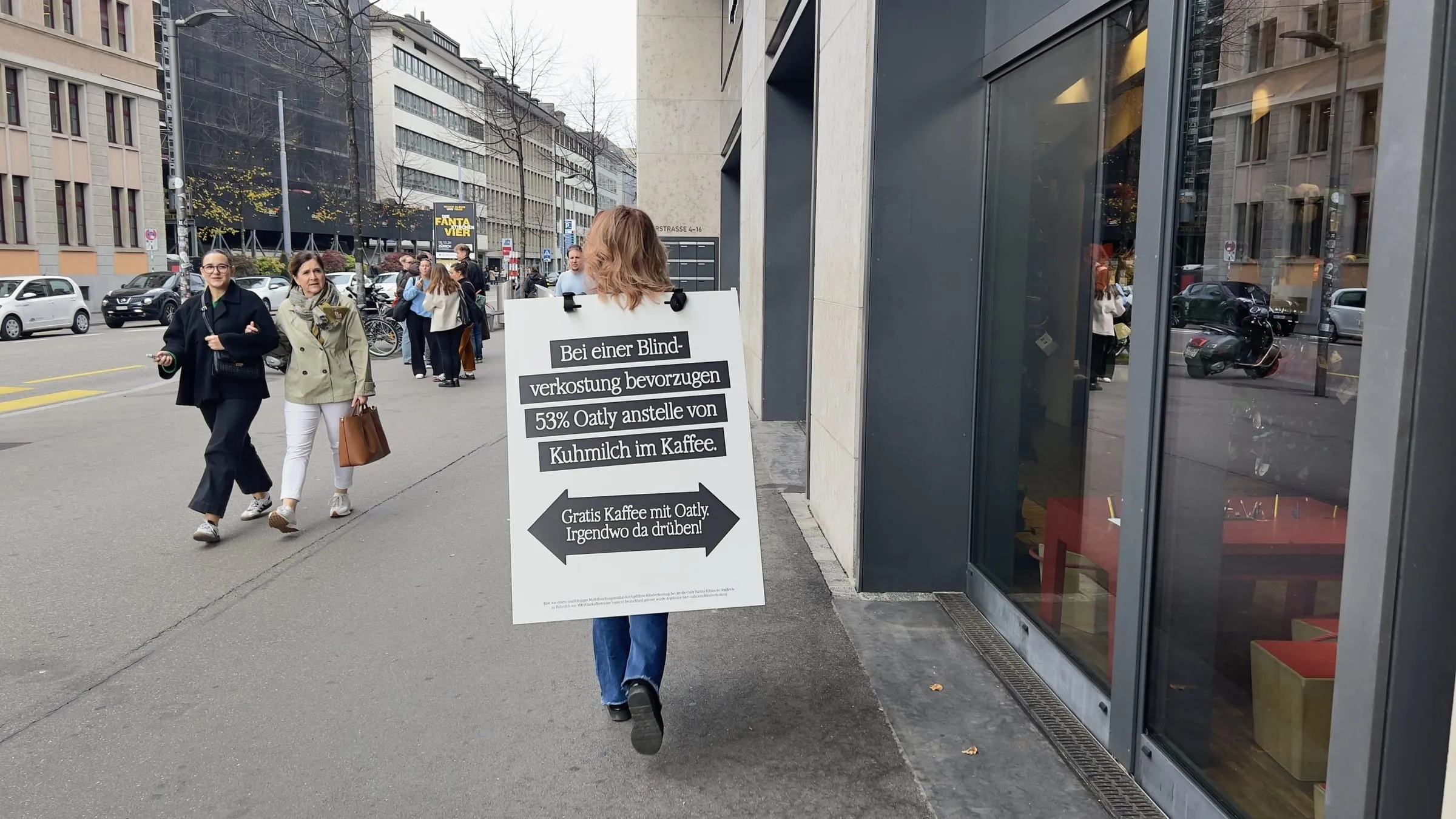 Sandwichfrau mit Oatly-Schild von hinten auf dem Trottoir in der Europaallee, entgegenkommende Passantinnen