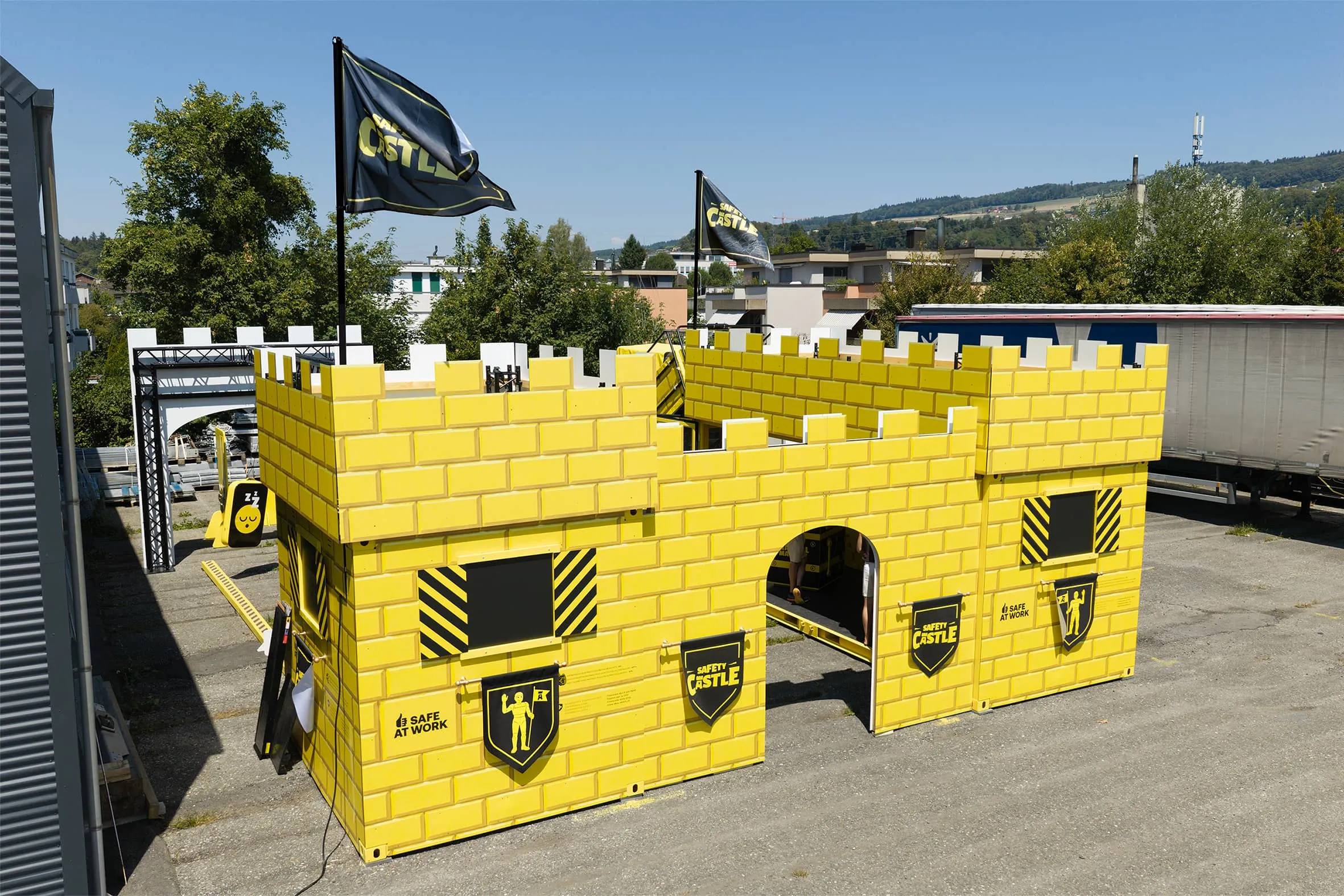 Drone shot of the yellow container castle with battlements, an arched gate, two flags and the Safety Castle crest on an outdoor site