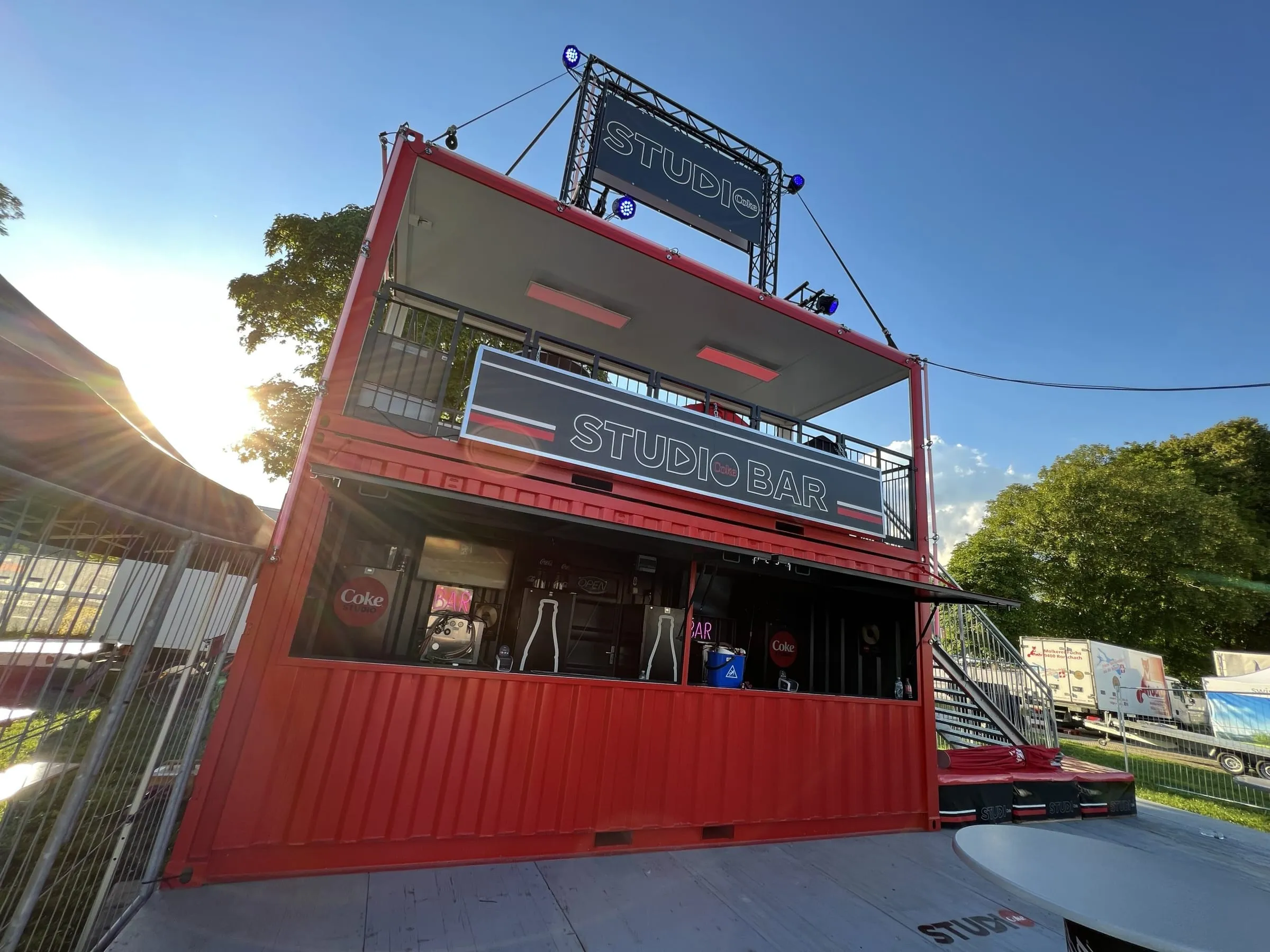 Red two-storey Coke Studio bar container with a balcony, truss structure and stairs, seen against the light