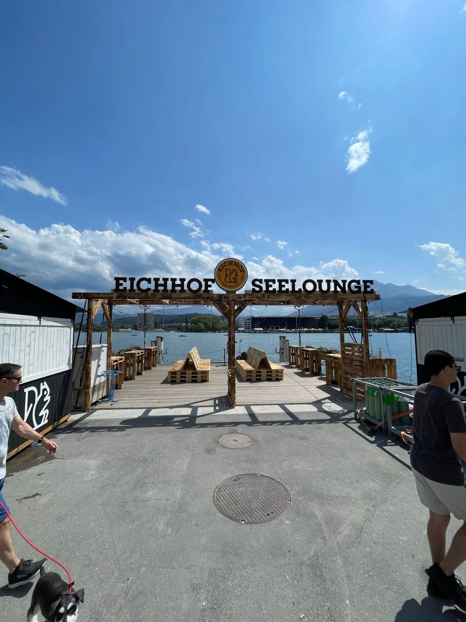 Pergola mit Schriftzug Eichhof Seelounge und Palettenmöbeln auf der Plattform am See, Berge im Hintergrund