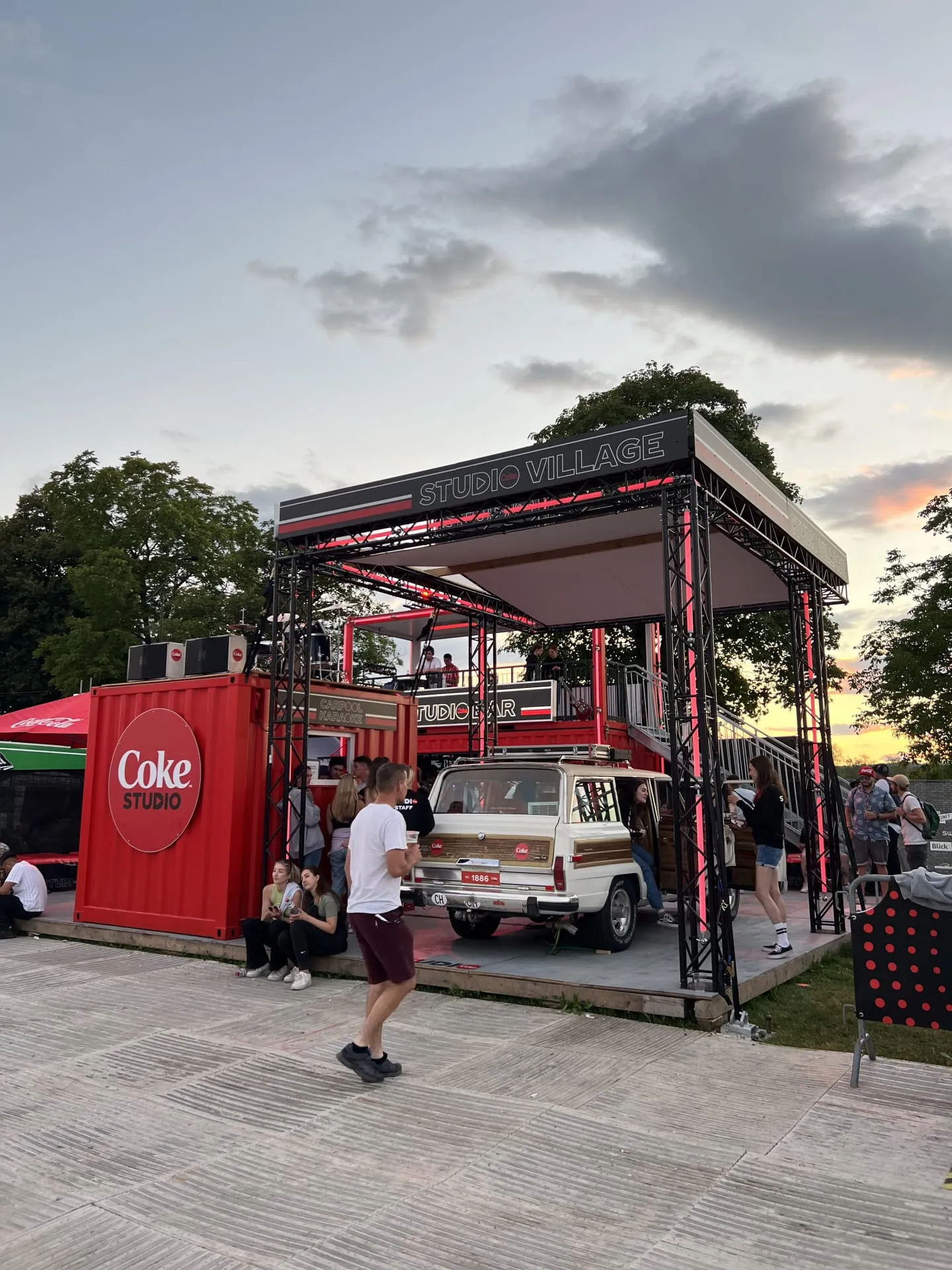 Roter Coke-Studio-Container mit Carpool-Karaoke-Jeep und Terrasse unter dem Studio-Village-Traversendach in der Abenddämmerung