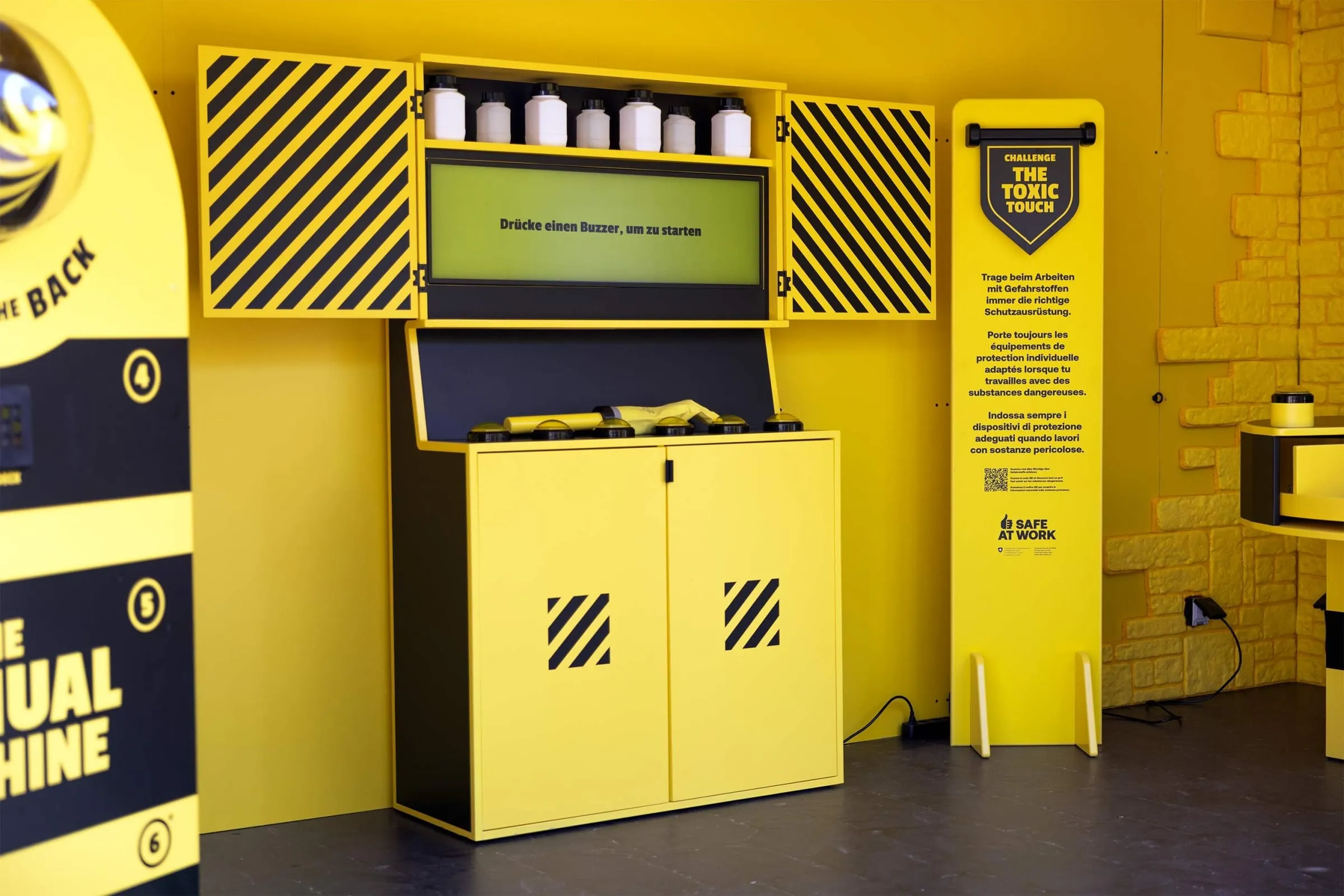 The Toxic Touch station: yellow hazardous-substances cabinet with white bottles, a screen reading «Drücke einen Buzzer, um zu starten» and a row of buzzers