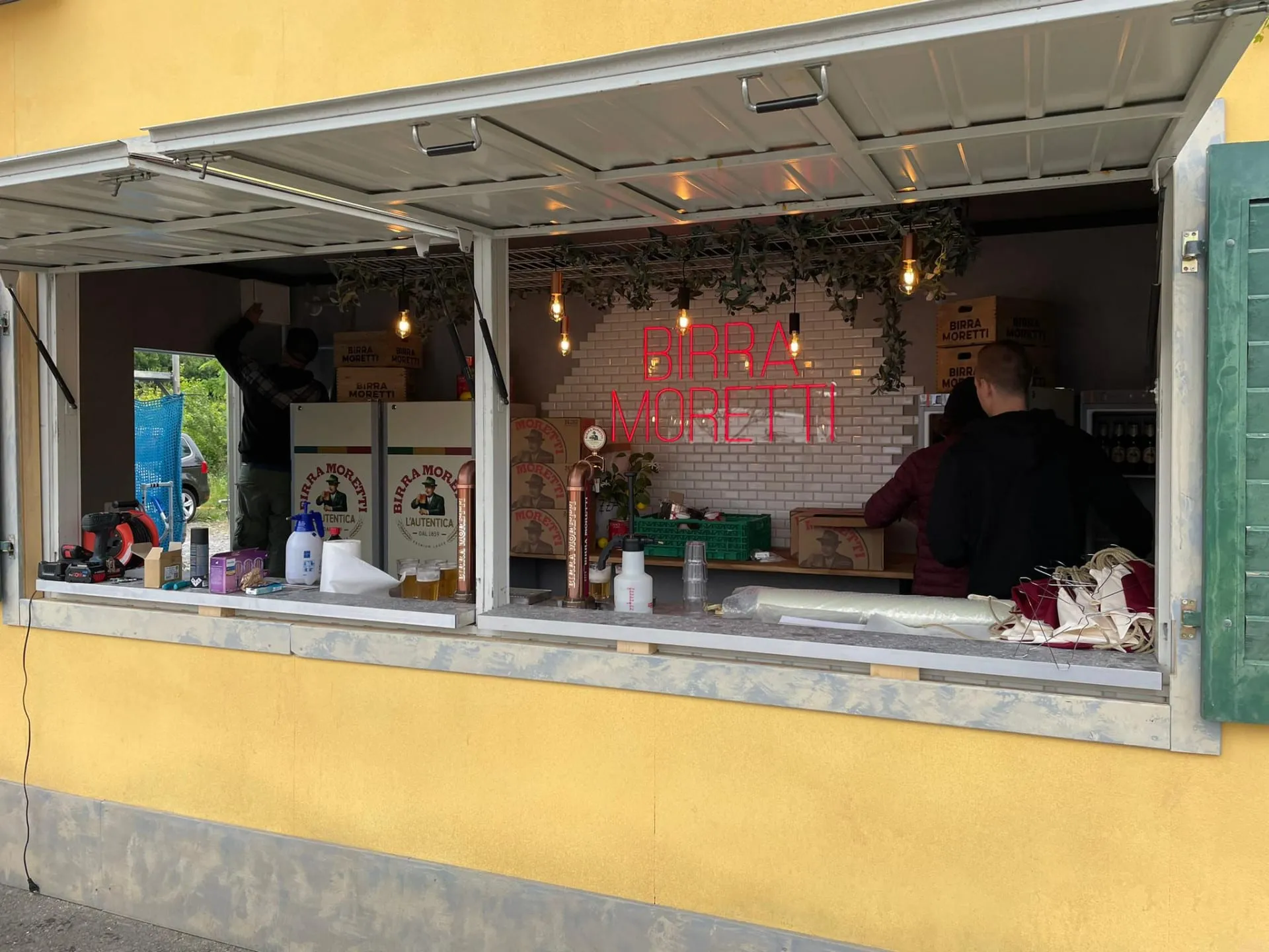 Offene Ausschankklappe der Container-Bar mit Neonschriftzug, Zapfhähnen und Team beim Einrichten