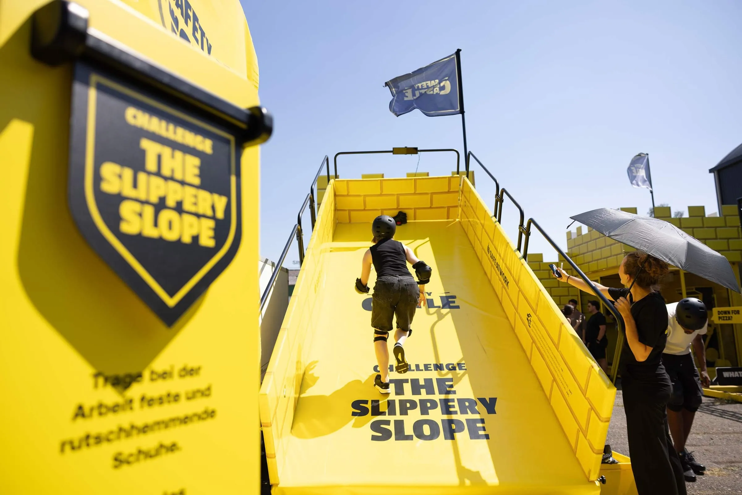 Young woman in a helmet and pads running up the yellow «The Slippery Slope» slide, Safety Castle flag in the background