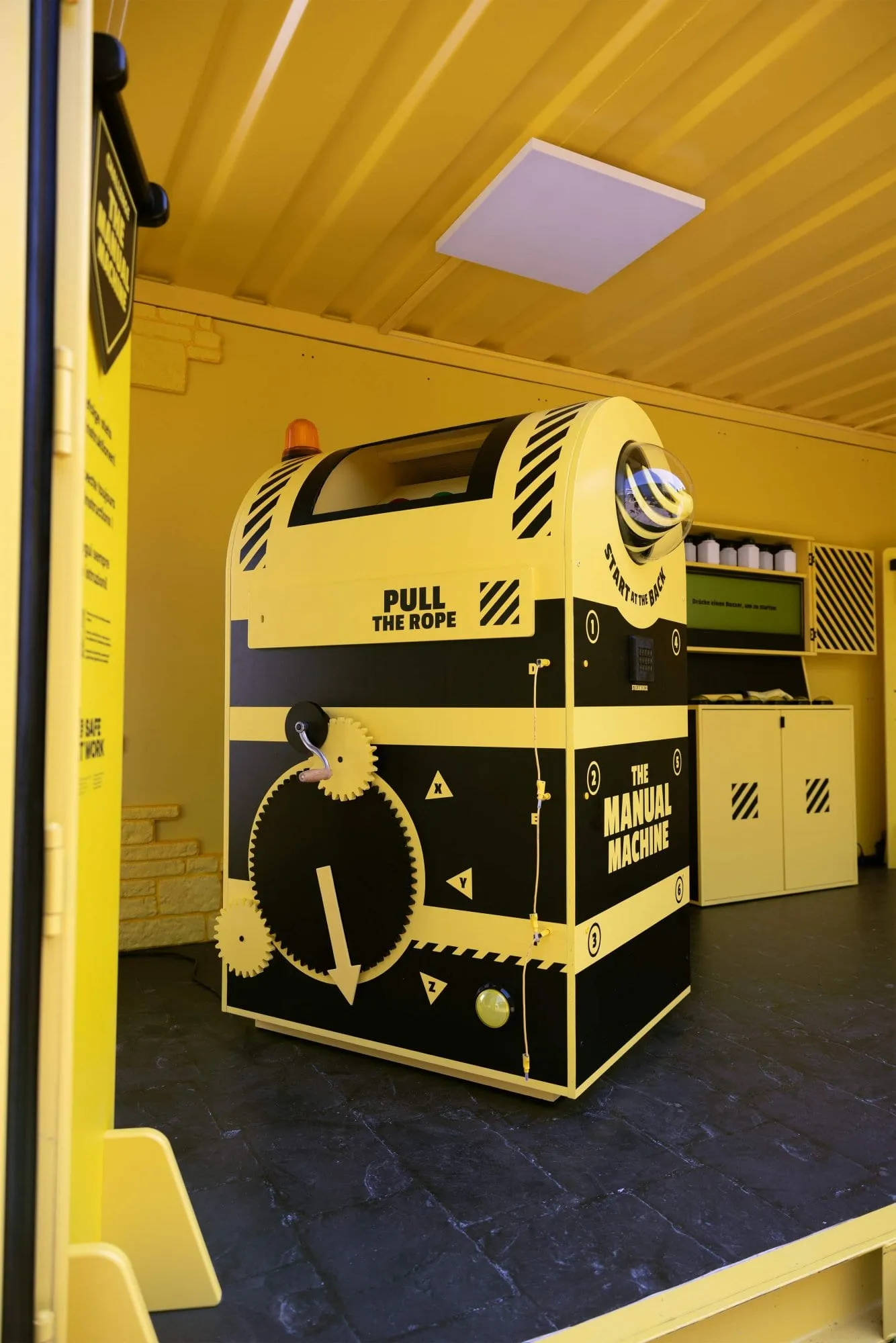 Yellow and black «The Manual Machine» station with a crank, gears, a warning light, a green buzzer and the words «Pull the Rope»