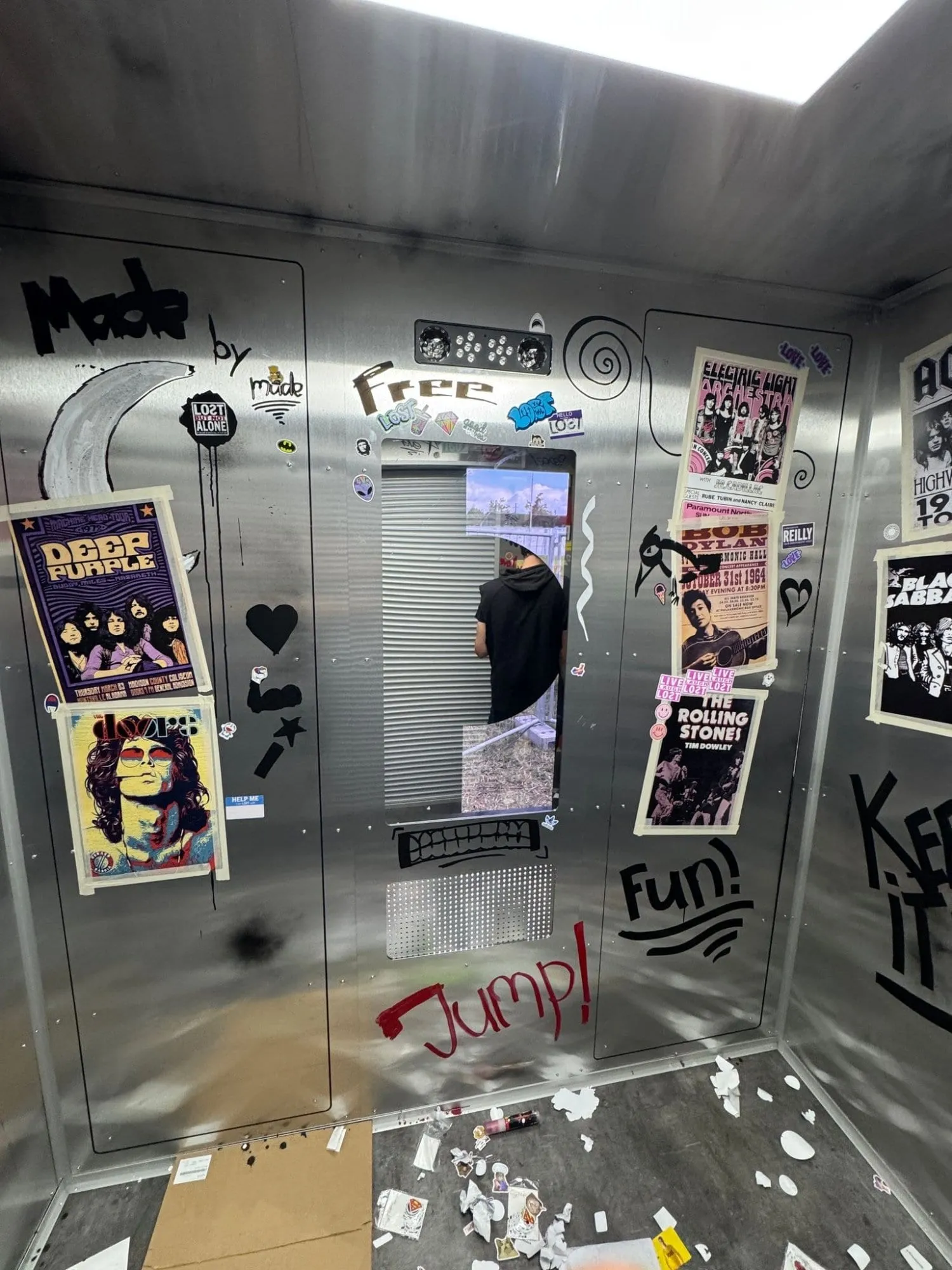 Innenraum im Lift-Look mit Edelstahlwänden, Konzertpostern, Stickern, Knopfleiste und Blick durch die Rolltor-Öffnung