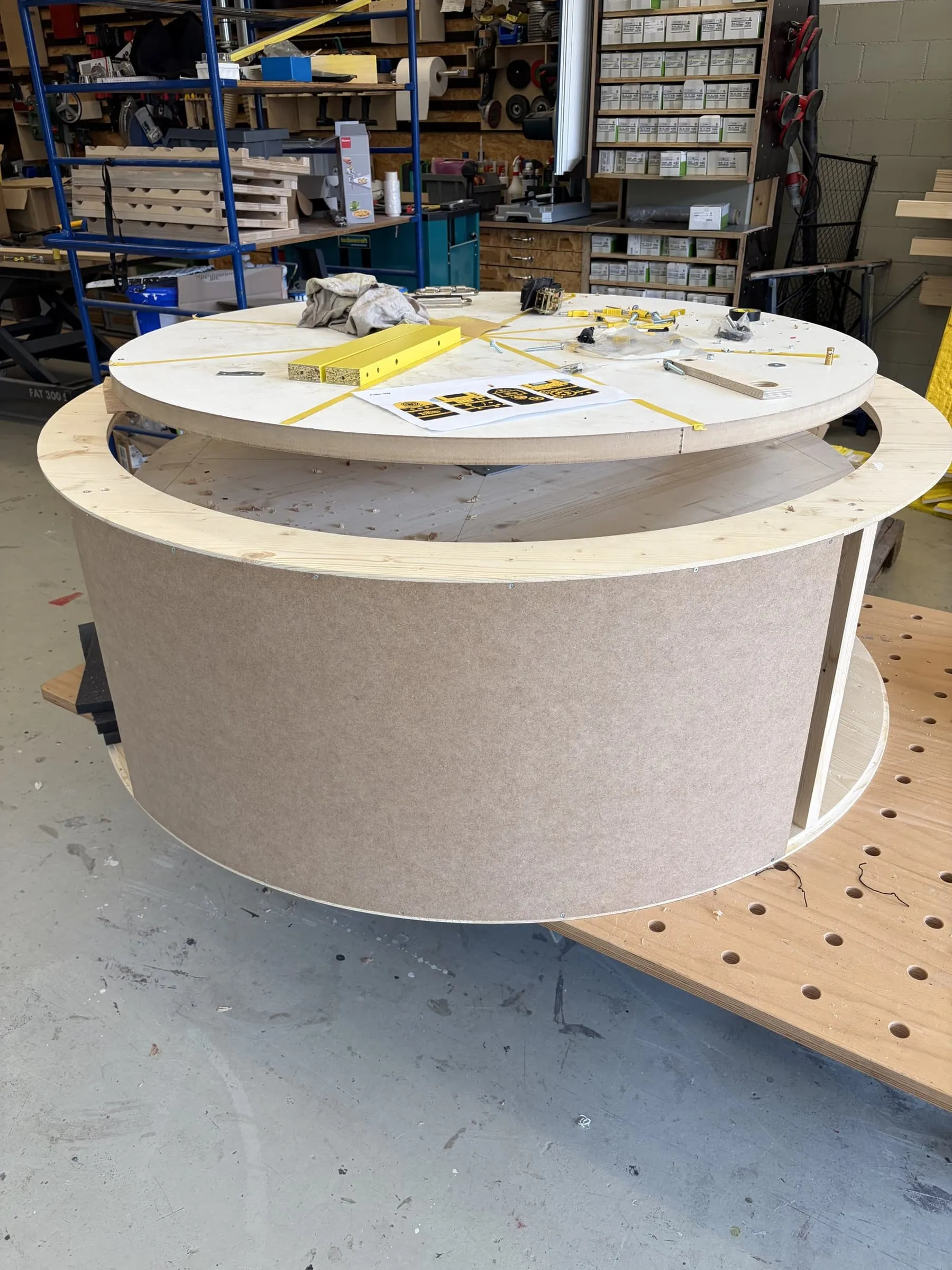 Unfinished round rotating platform made of wood and MDF on the workshop table, with screws, yellow battens and a printout on it
