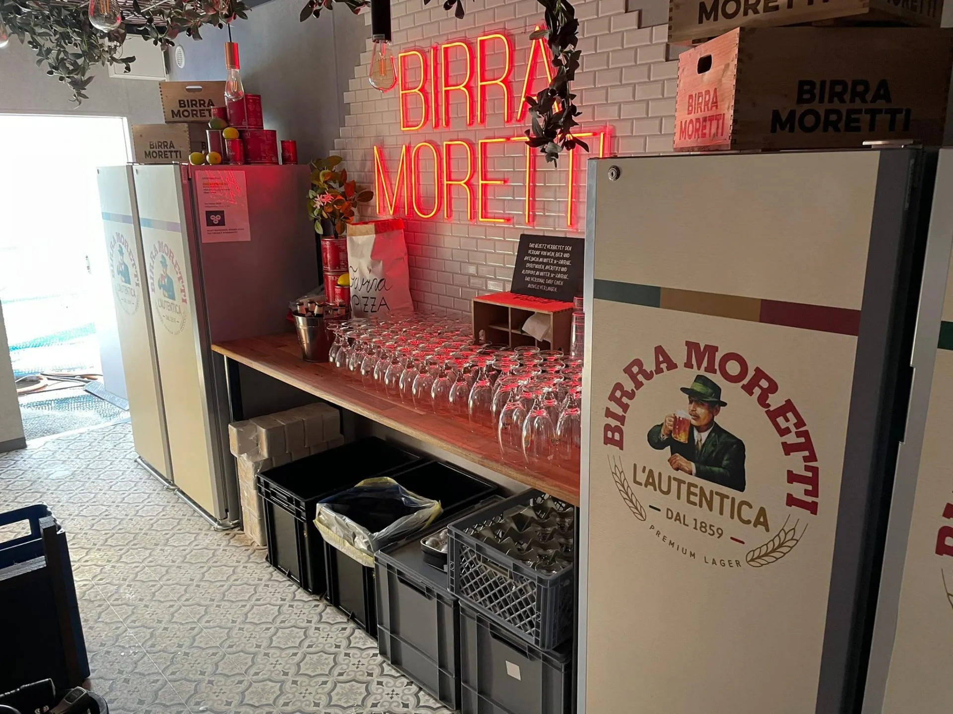 Container-Bar innen mit weissen Fliesen, rotem Neonschriftzug Birra Moretti, Gläsern und gebrandeten Kühlschränken