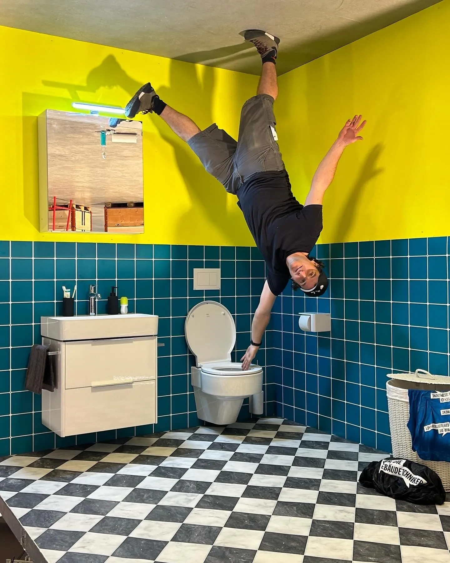 Mann hängt kopfüber im Upside-Down-Bad mit gelben Wänden, blauen Plättli und Schachbrettboden