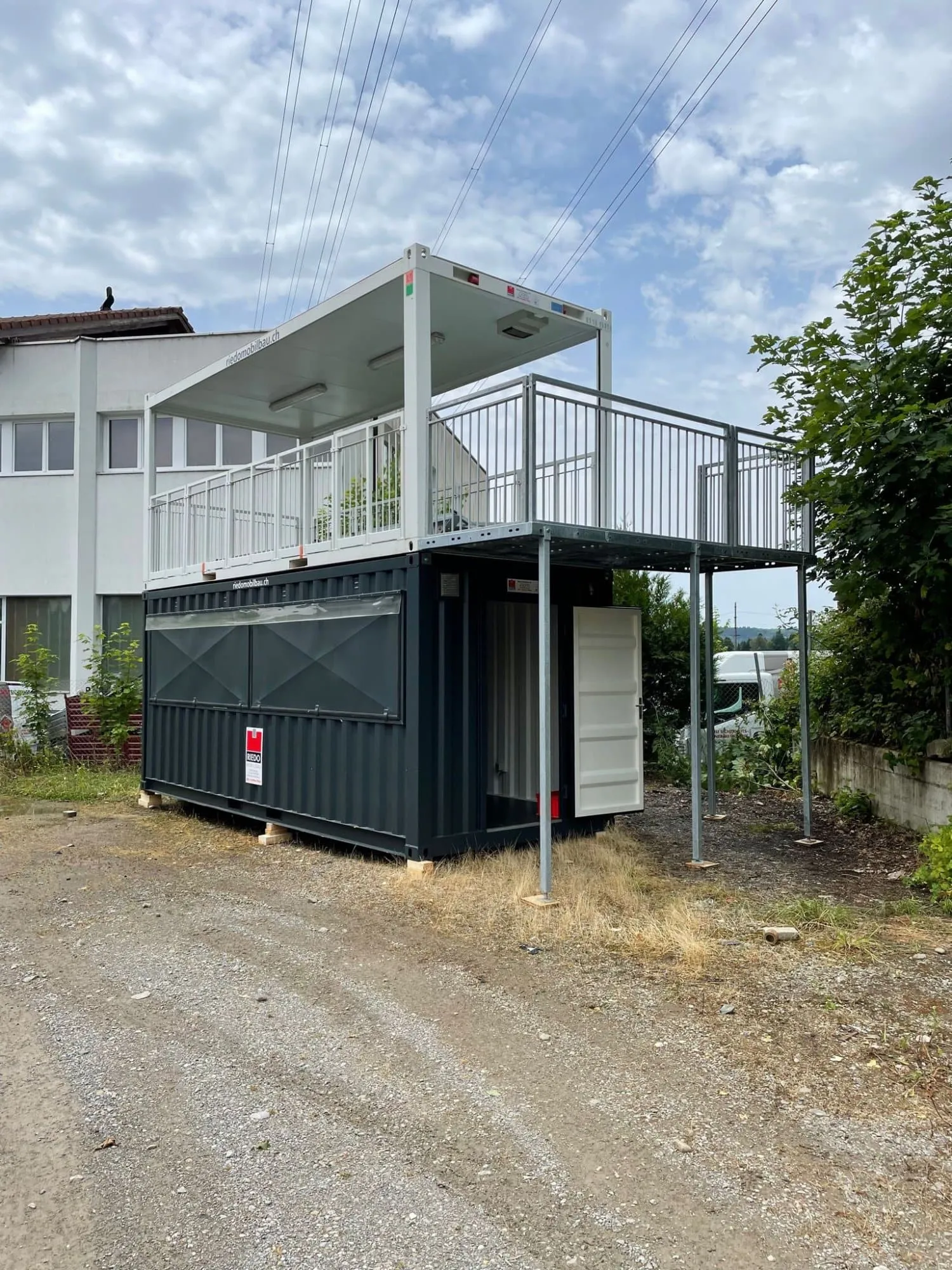 Anthrazitfarbener Container mit Dachterrasse und Geländer auf Stützen vor dem Umbau