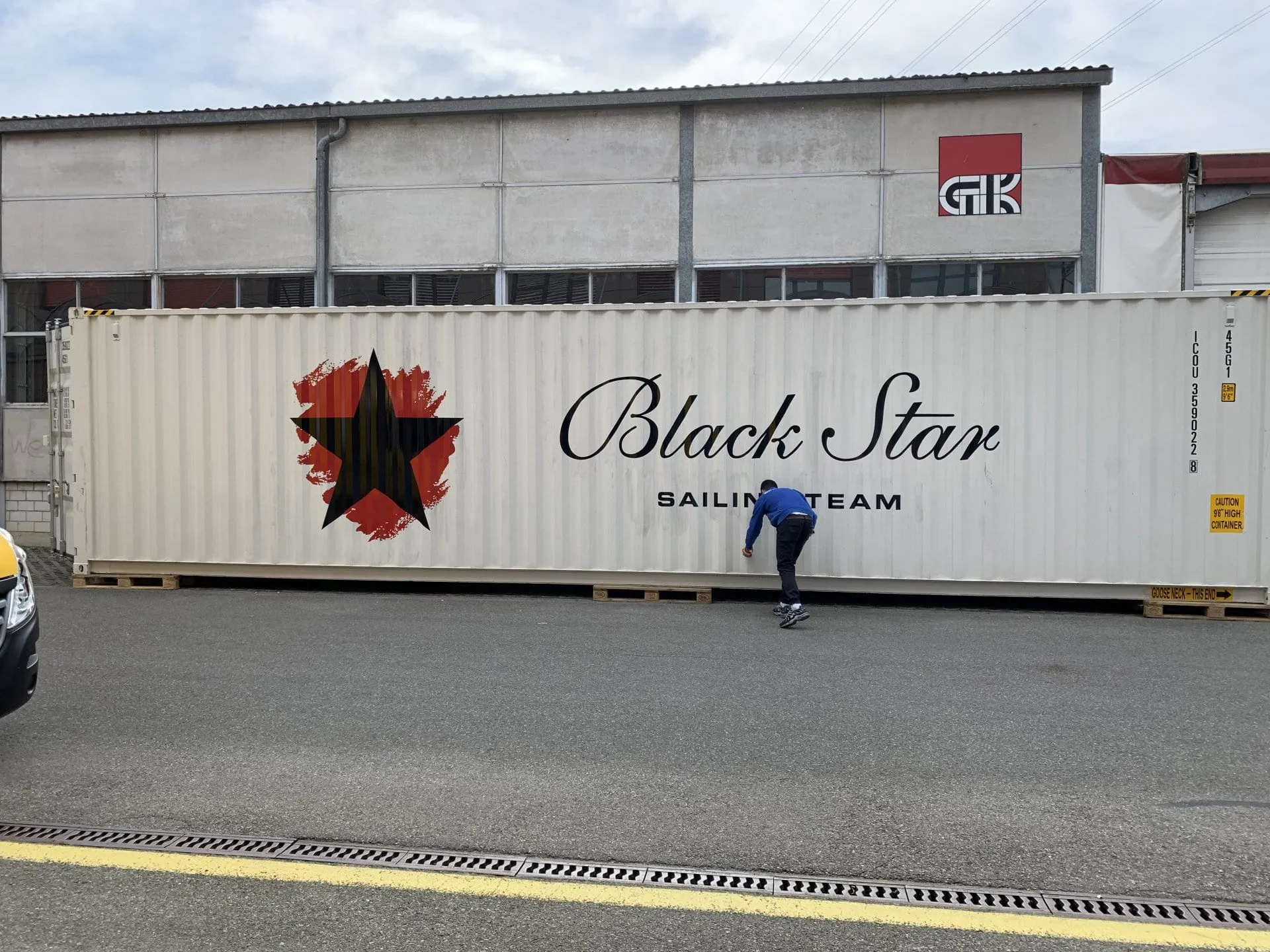 Weisser Container mit rotem Stern und Schriftzug «Black Star Sailing Team» vor einer Werkhalle, ein Mann beim Anbringen
