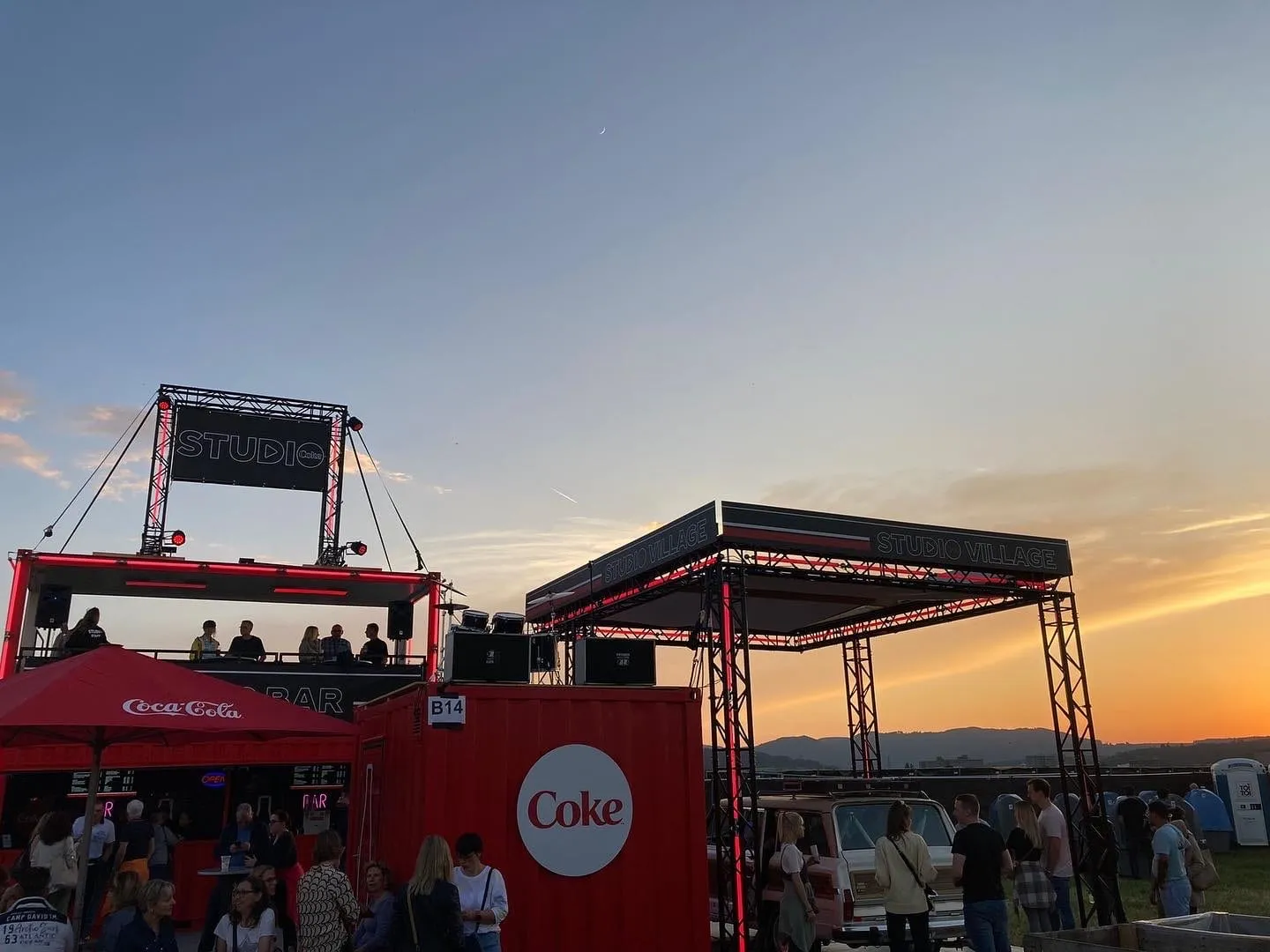 Gesamter Coke-Studio-Stand mit Bar, Terrasse, rotem Container und Traversen im Sonnenuntergang, davor Besuchende