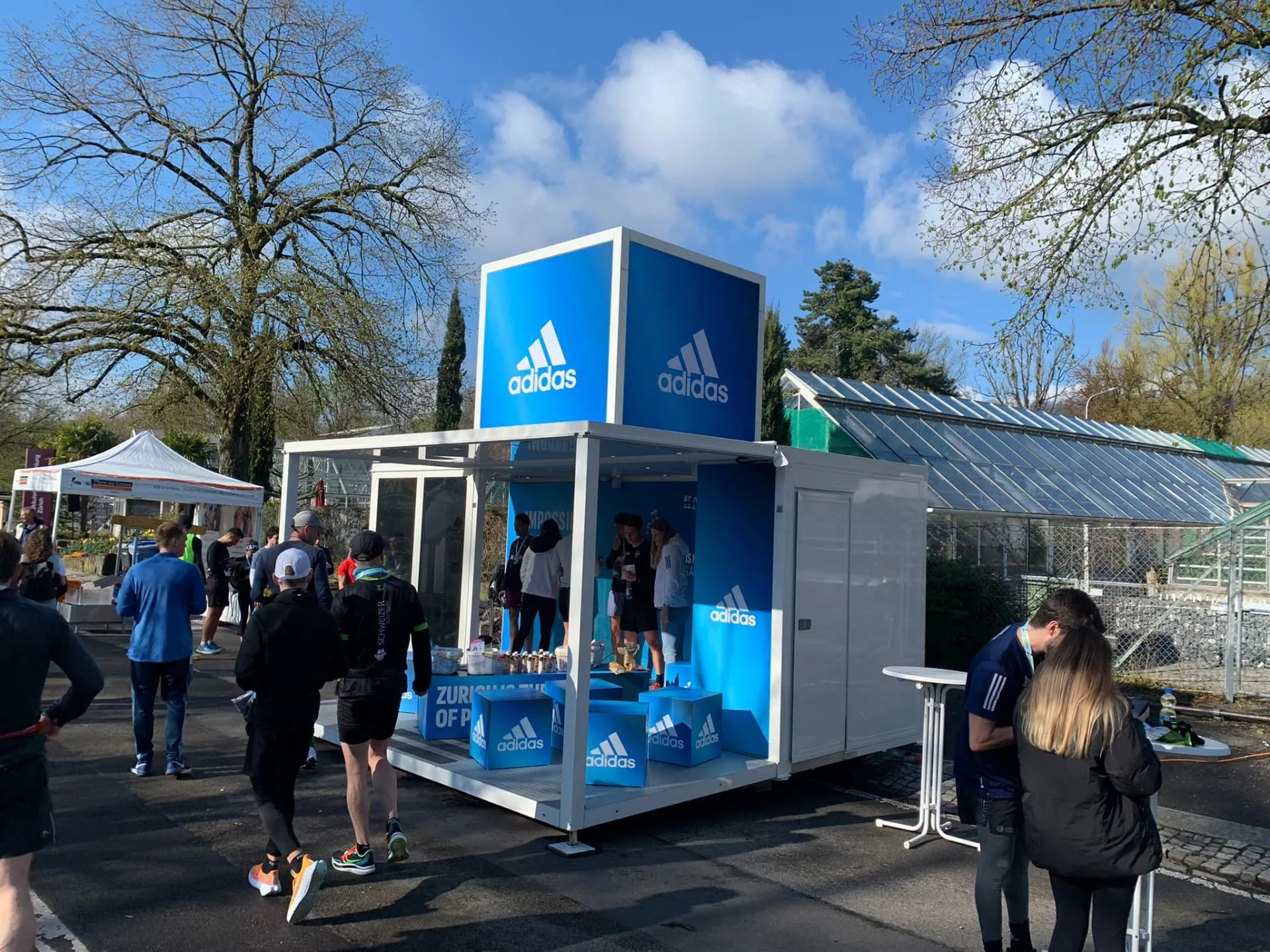 Blau-weisser adidas-Promocube mit Würfel auf dem Dach, davor Läufer und Besuchende bei einem Stehtisch