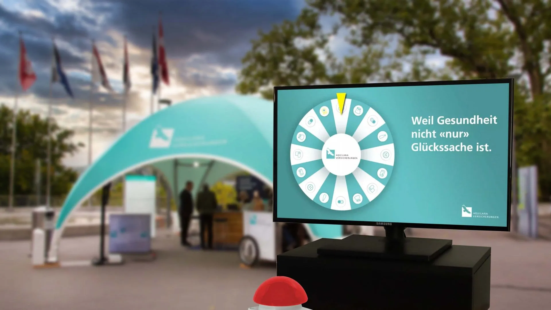 Screen with the turquoise Aquilana Glücksrad and a red buzzer, a turquoise tent with flags behind it