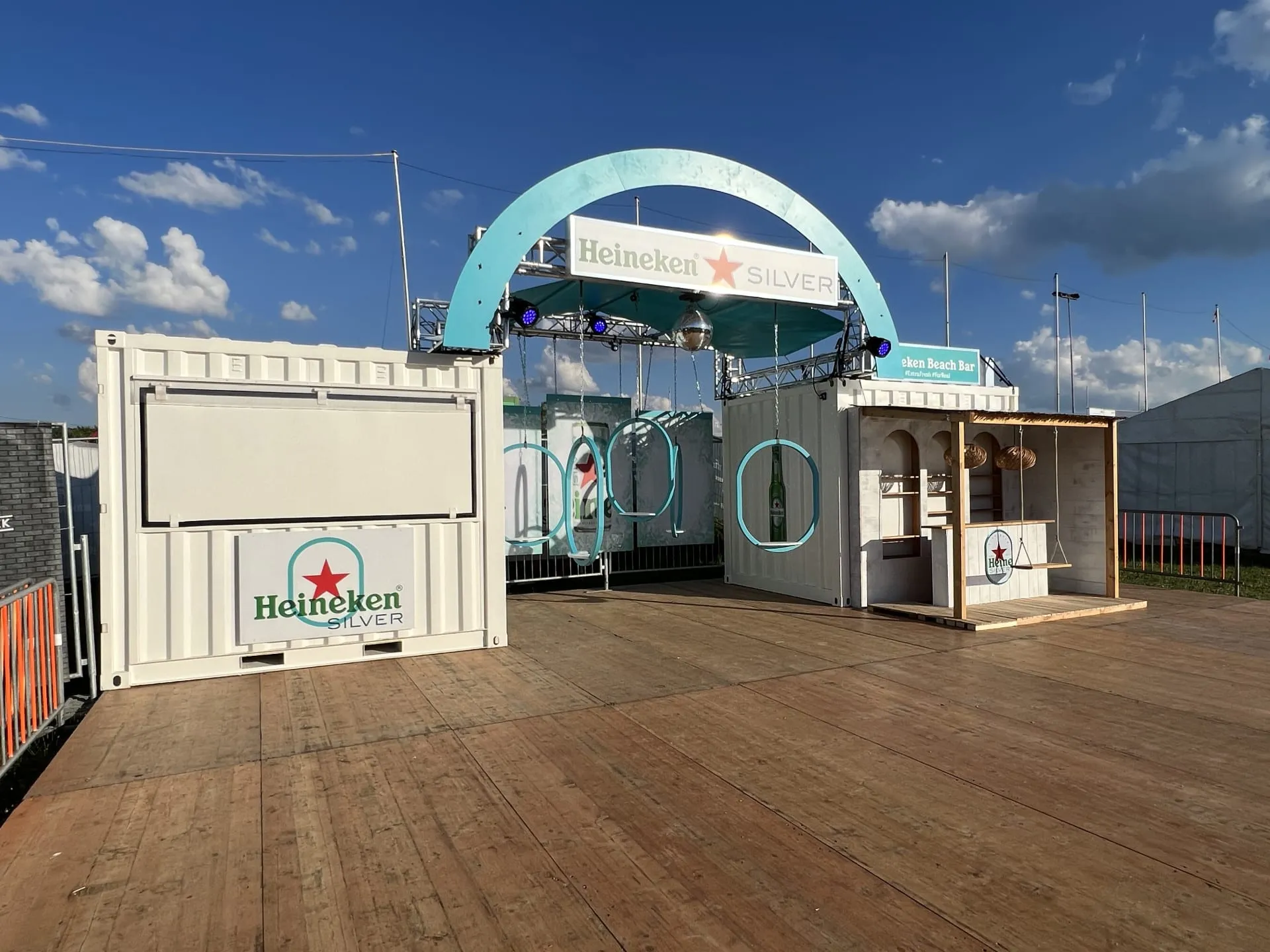 Weisse Heineken-Silver-Container mit türkisem Bogen, Ringschaukeln und Beach Bar auf Holzpodest vor dem Festival