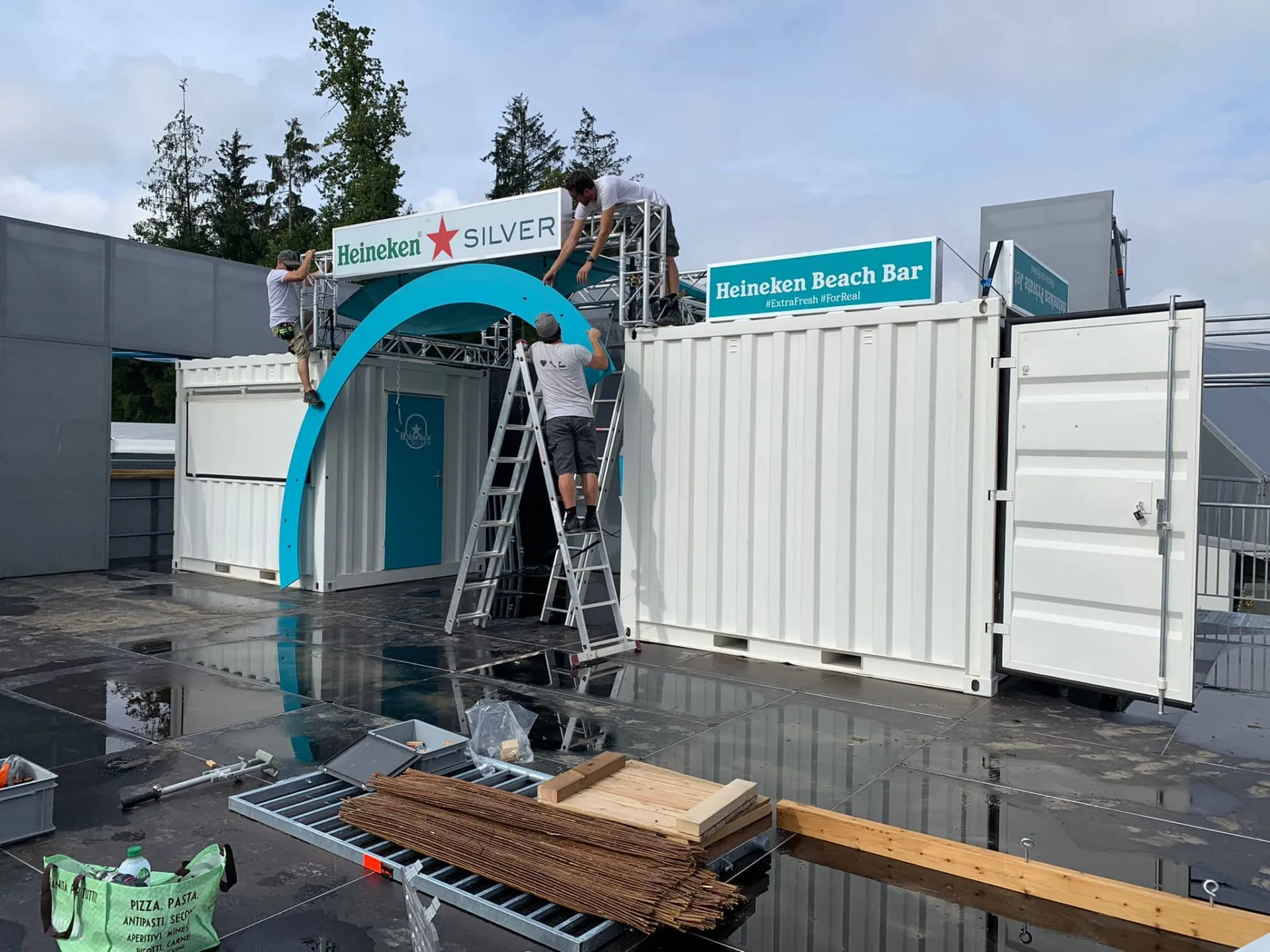 Drei Männer montieren Bogen und Schild auf den Traversen über den weissen Containern, Bauteile am Boden