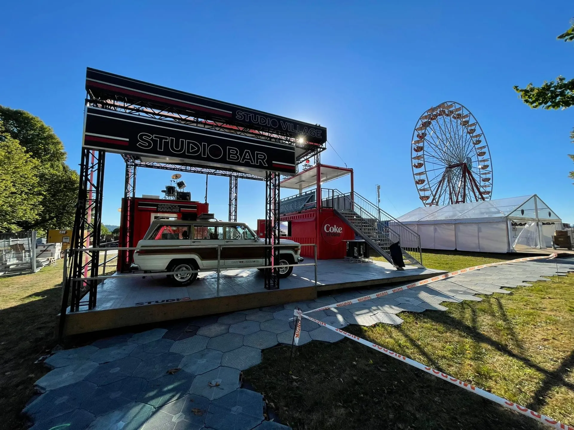 Weisser Jeep Wagoneer unter dem Studio-Bar-Traversendach neben dem roten Container, dahinter Riesenrad und Zelt