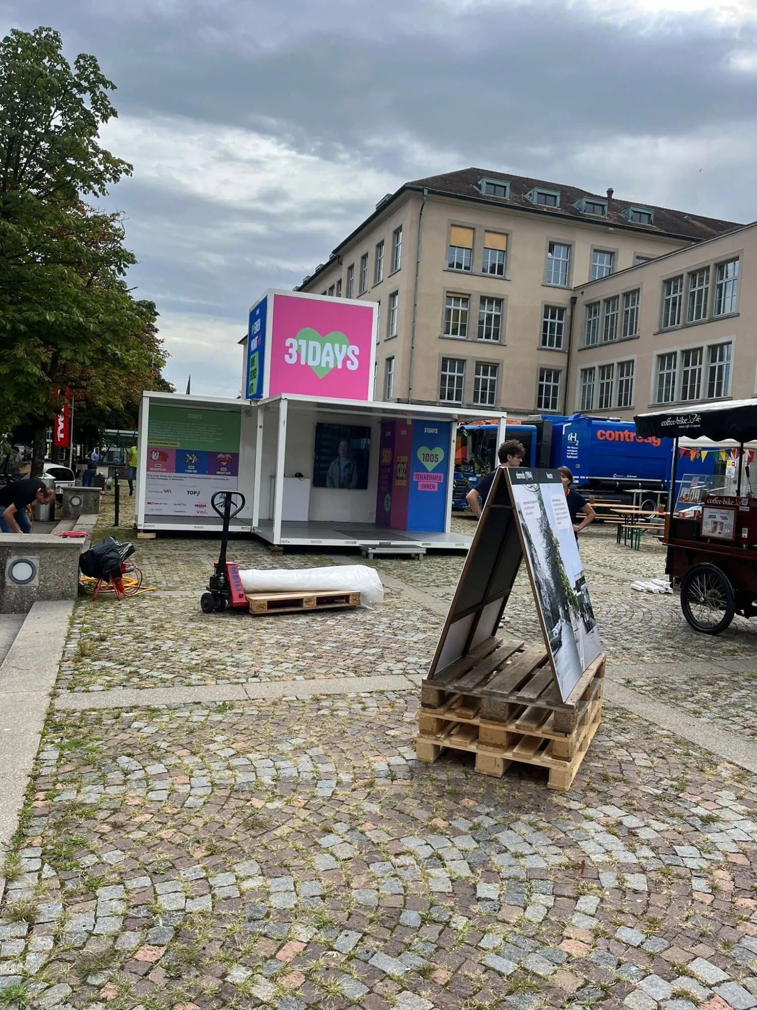 Aufbau des Promocubes mit Hubwagen, Palette und Kundenstopper auf dem Pflasterplatz vor einem Schulgebäude