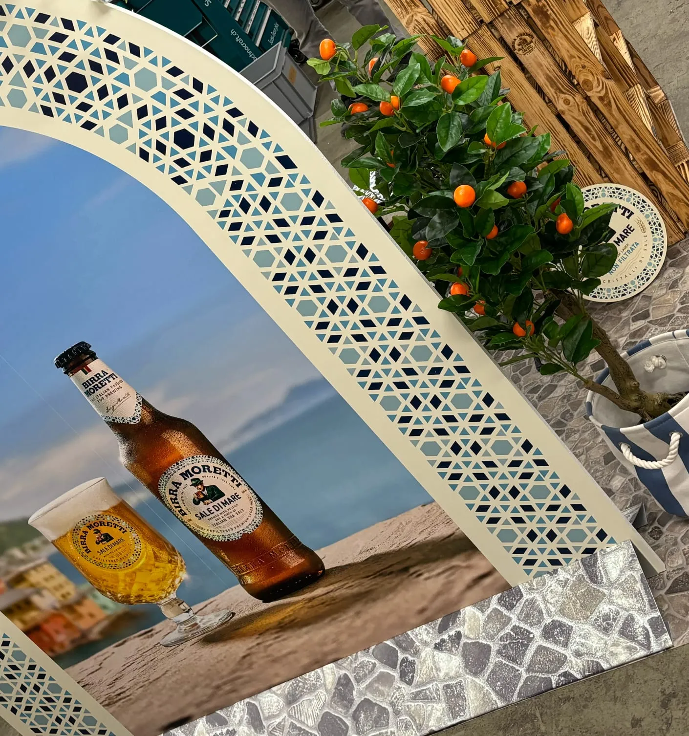 Detail des Torbogens mit blau-weissem Mosaikmuster, Bierflasche und Glas auf dem Motiv, daneben ein Orangenbäumchen