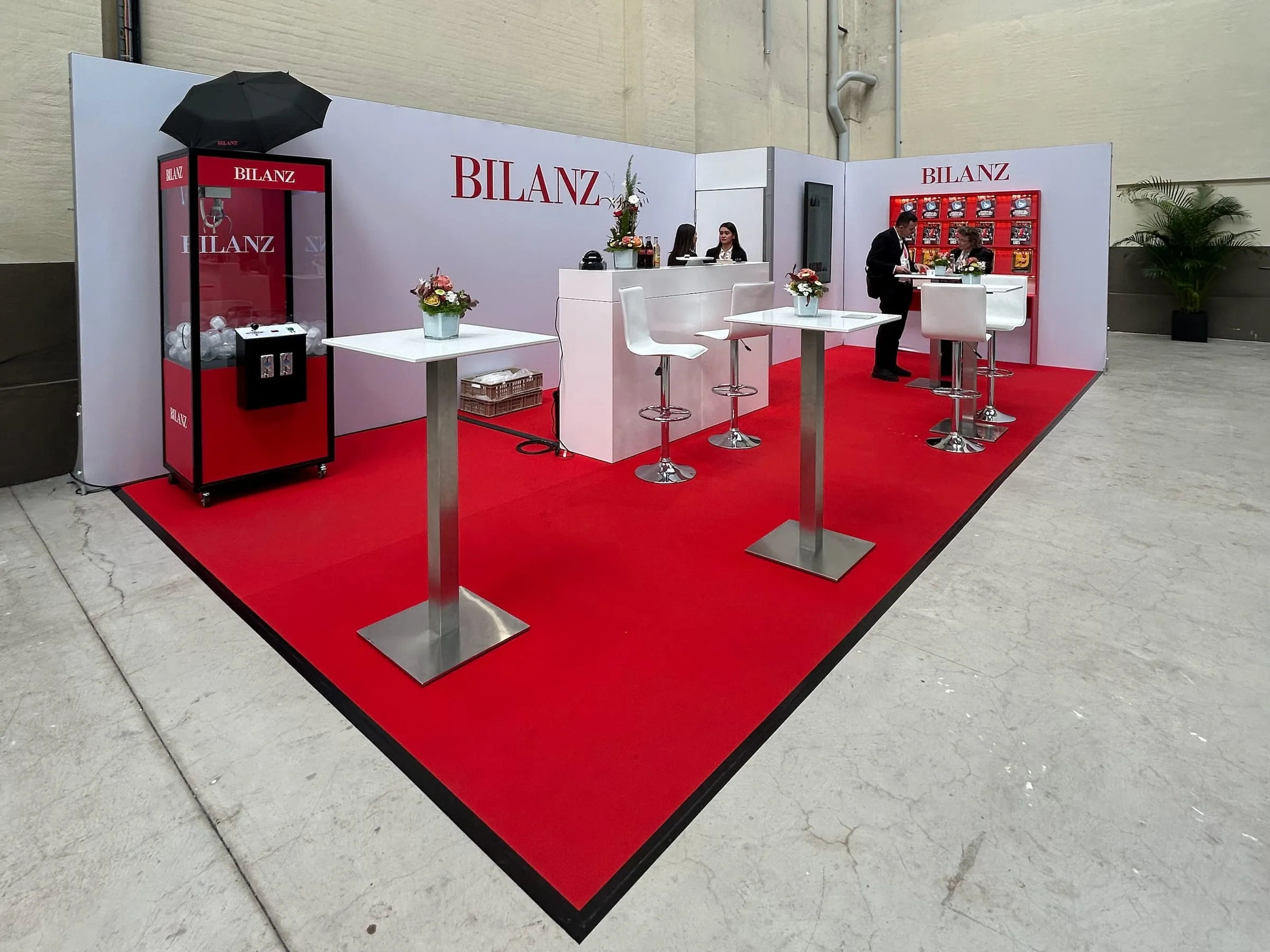 Übersicht des BILANZ-Stands mit rotem Teppich, weissen Stehtischen, Bar, Greifarmautomat und Magazinregal in der Halle 550