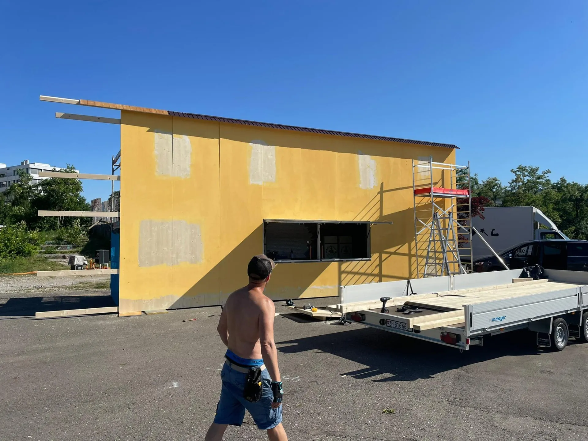 Gelbe Fassade mit ausgesparten Fensterflächen und Gerüst vor Ort, davor Anhänger mit Holzbalken und Arbeiter