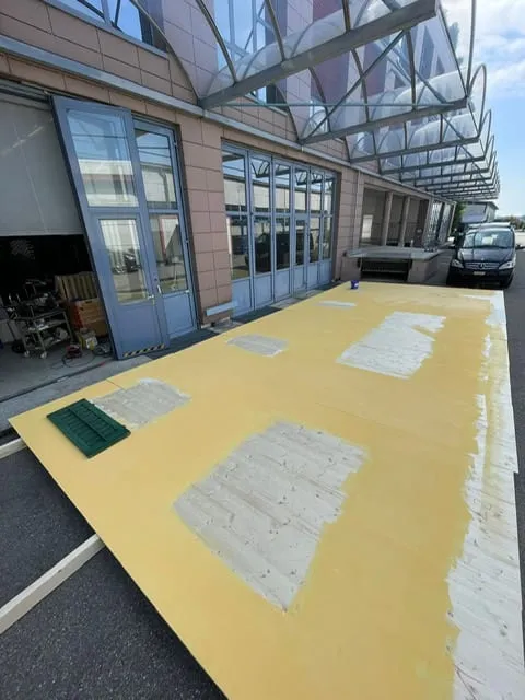 Gelb gestrichene Fassadenplatten mit ausgesparten Fensterflächen und grünem Fensterladen am Boden vor der Werkstatt