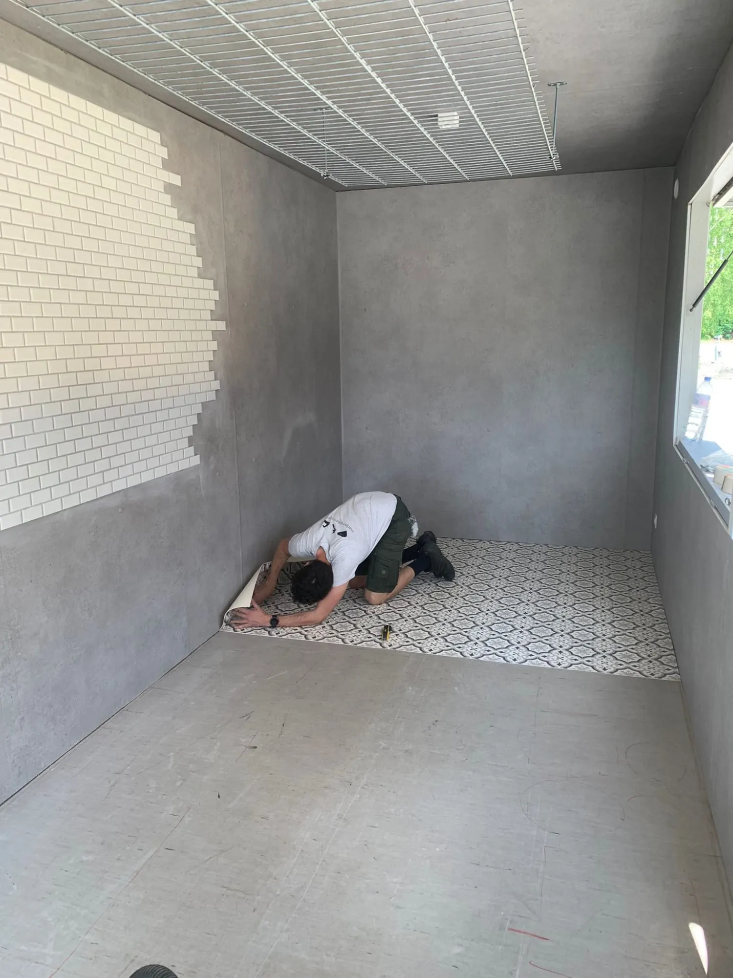 Mann verlegt gemusterten Vinylboden im leeren Container mit grauen Wänden und weissen Fliesen