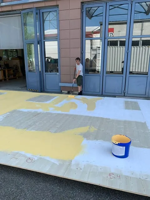 Mann streicht grosse Holzplatten mit gelber Farbe vor dem Werkstattgebäude, Farbkessel daneben