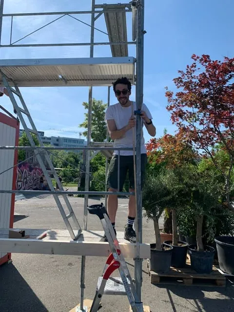 Mann mit Sonnenbrille steht auf dem Gerüst neben dem Container, im Hintergrund Topfbäume