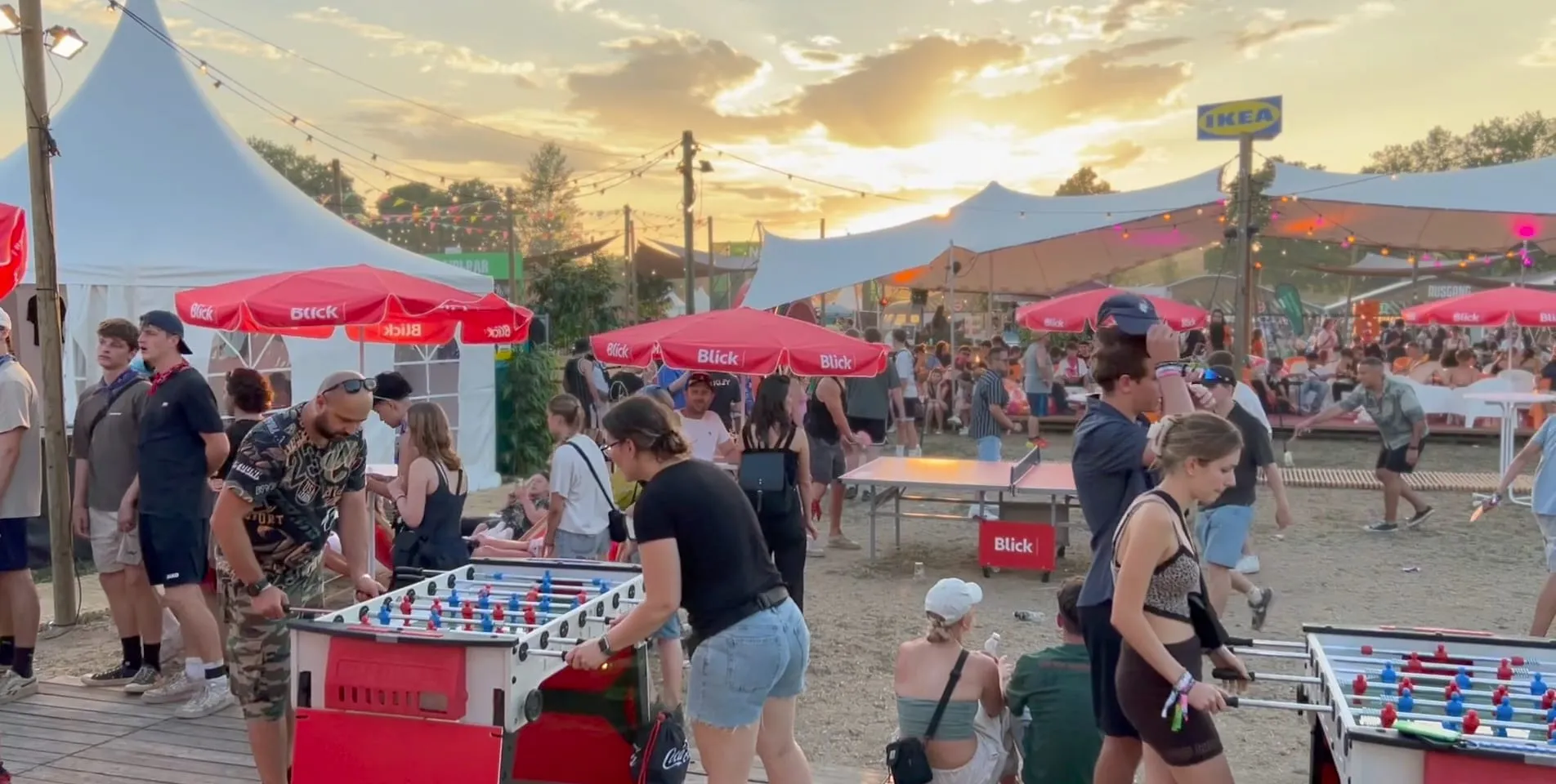Festival visitors playing table football under red Blick parasols, with a table-tennis table and tents behind them in the evening light