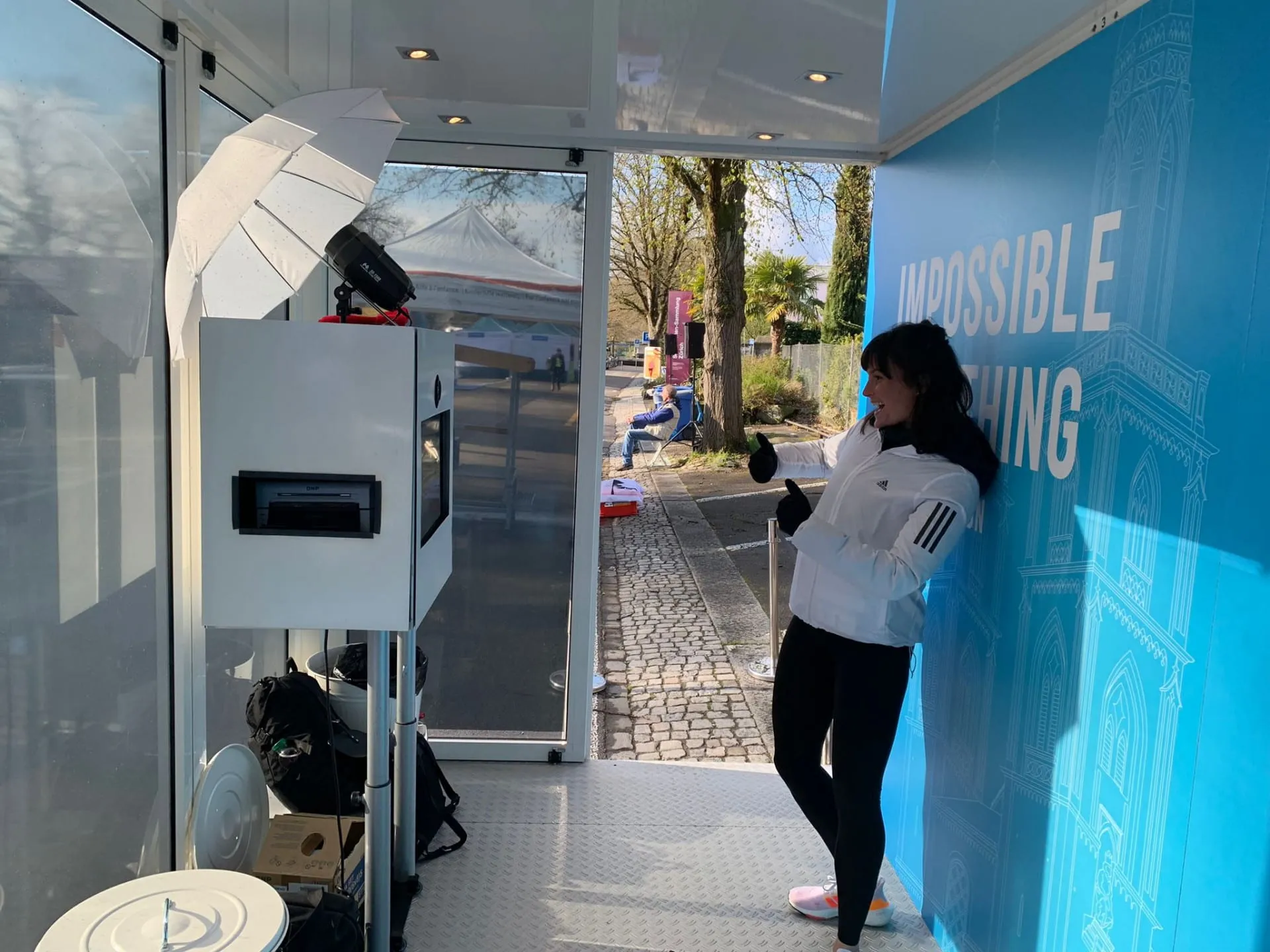 Frau in weisser adidas-Jacke posiert mit Daumen hoch vor der Fotobox mit Blitzschirm im Promocube