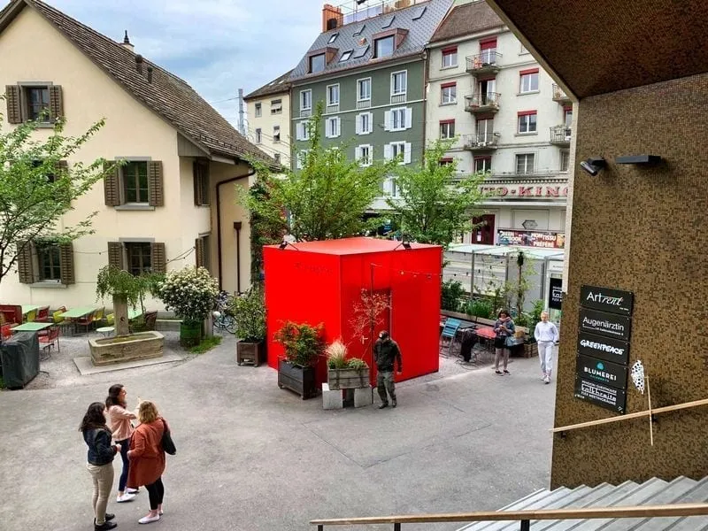 Red cube container on a courtyard between apartment buildings, passers-by in front of it