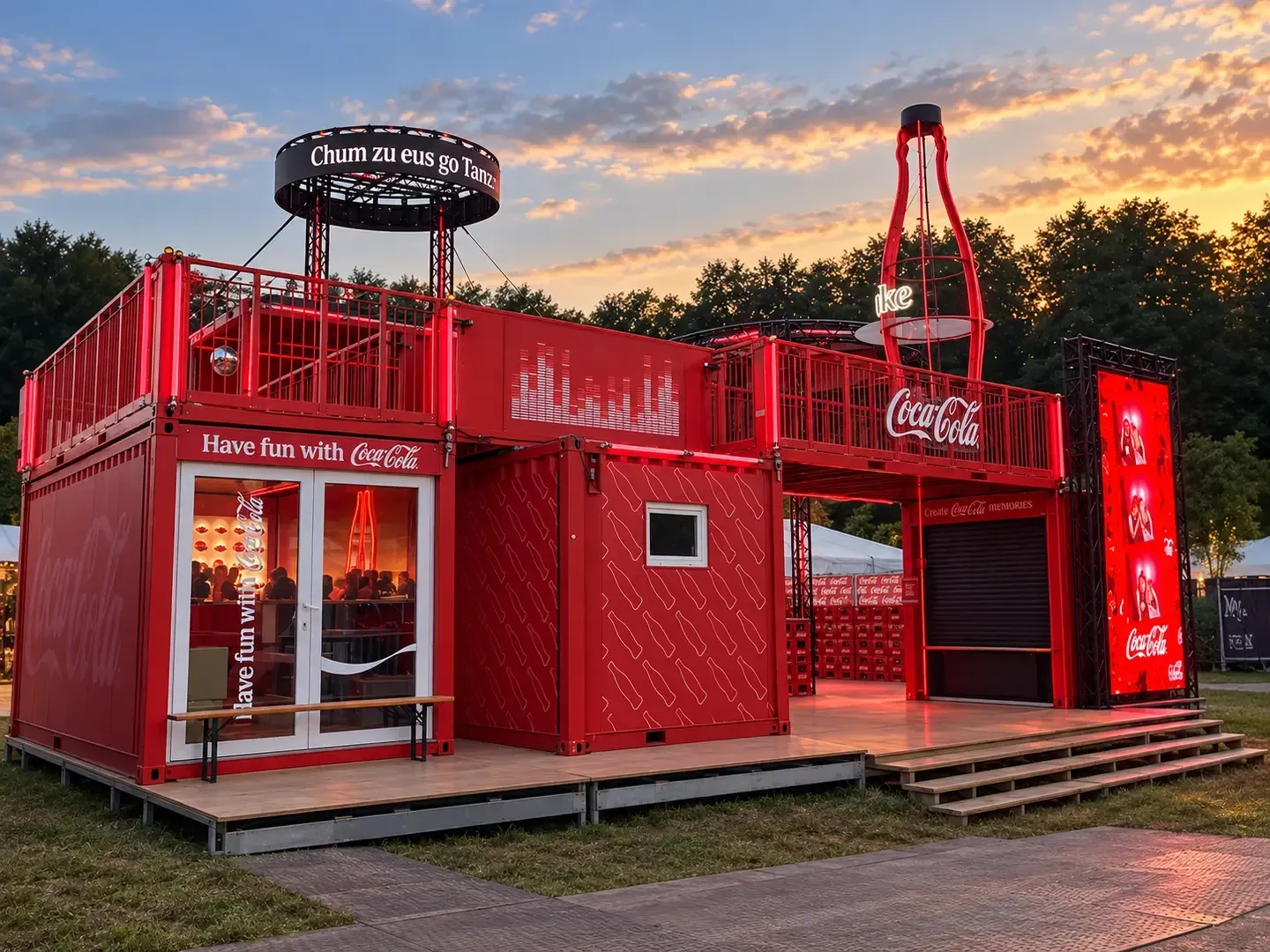 Roter Coca-Cola-Container am Festival in der Abenddämmerung: zwei gestapelte Container mit Dachterrasse, leuchtender Flaschen-Skulptur, Bar und Screen, drinnen Gäste