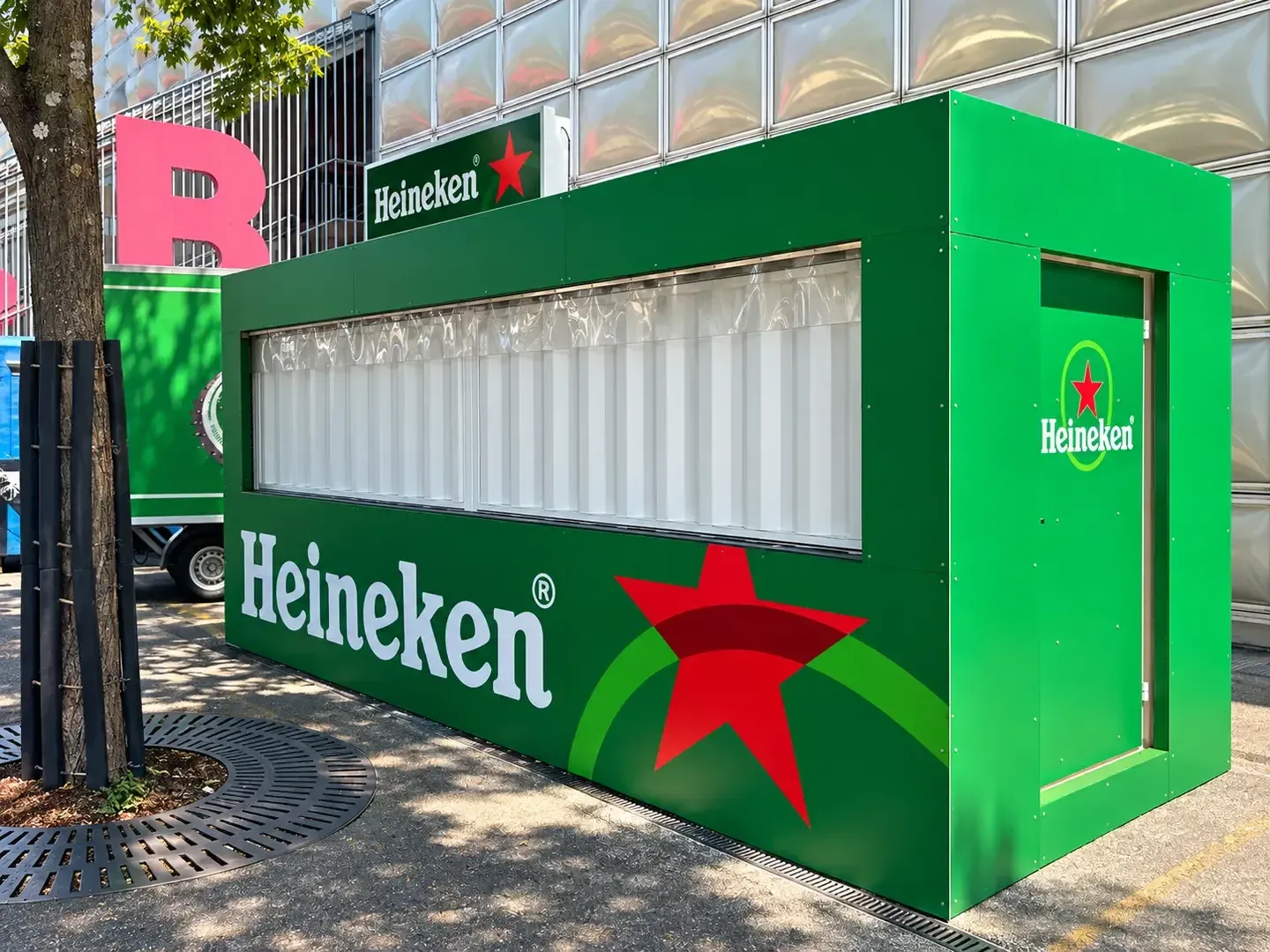 Grüner Heineken-Barcontainer mit geschlossener Klappe und Leuchtkasten auf dem Dach vor einer Fassade aus Glasbausteinen