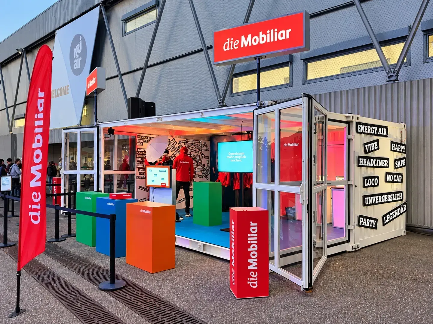 Weisser Container der Mobiliar mit verglaster Front vor einer Eventhalle, drinnen ein Screen und bunte Würfel, aussen Beachflag und Säulen in Rot