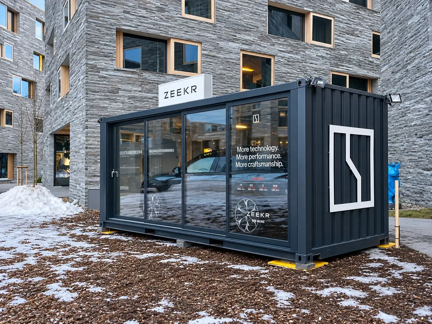 Anthrazitfarbener Zeekr-Container mit verglaster Längsseite und Schiebetüre vor Steinfassaden in Laax, drinnen ein Auto, Schneereste am Boden