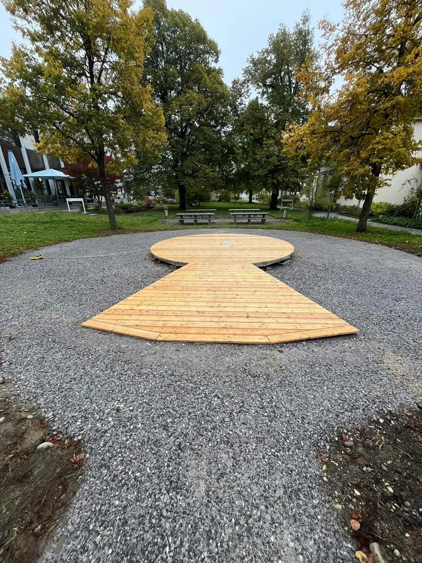 Rundes Holzdeck mit Rampe auf Kiesfläche im Park, dahinter Bäume in Herbstfarben