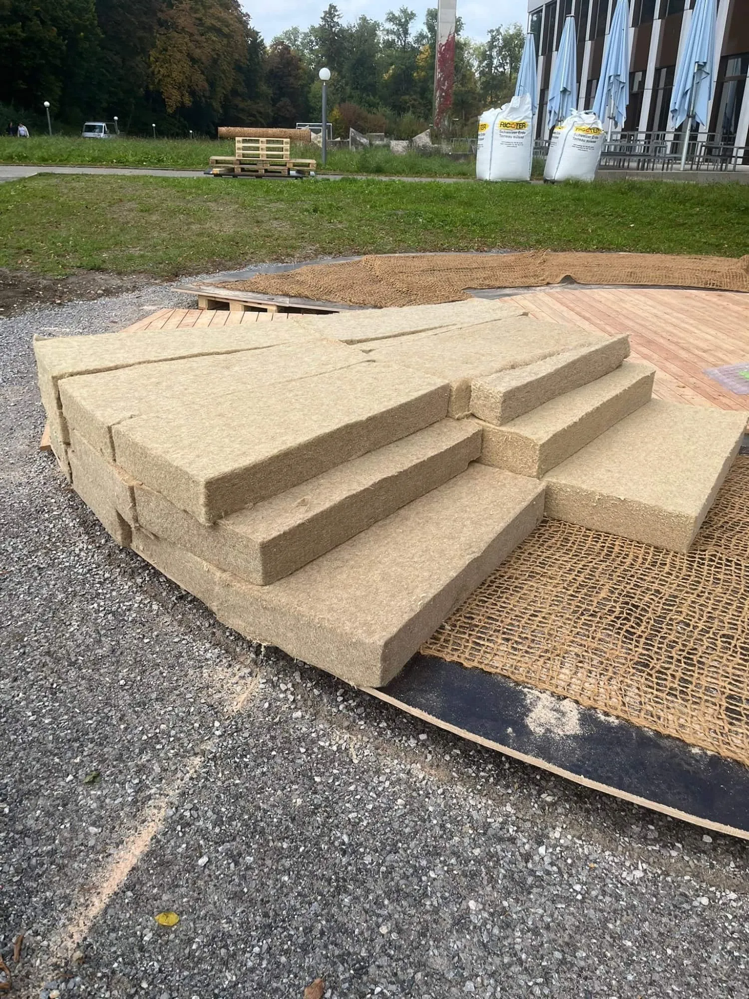 Gestapelte Hanf-Isoliermatten und Jutenetz am Rand des Holzdecks, dahinter Big Bags mit Erde