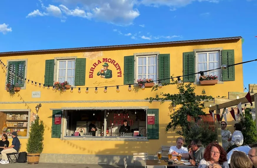 Yellow Birra Moretti building facade with green shutters, a mural, string lights and guests at the bar