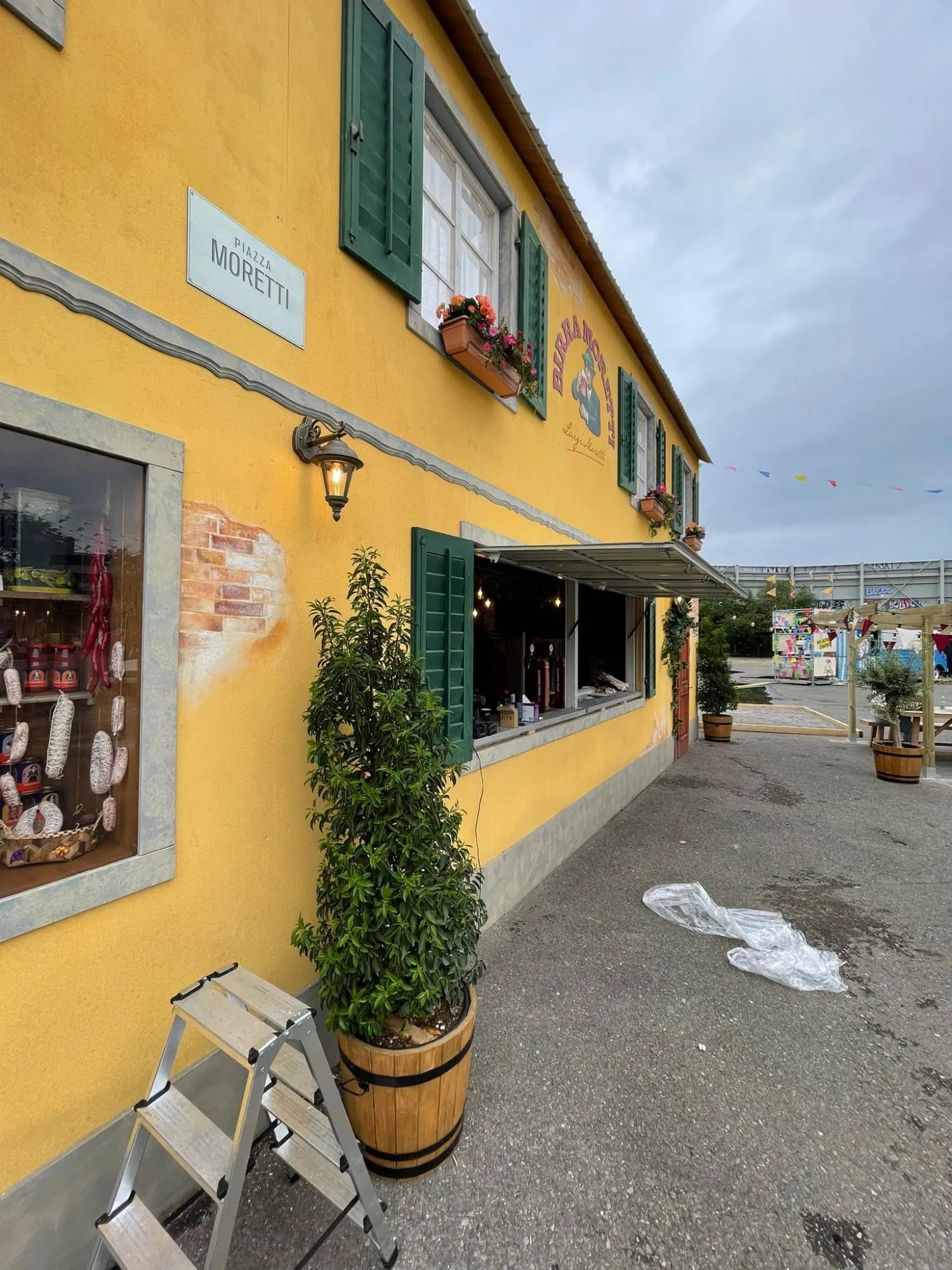 Seitenansicht der gelben Fassade mit Schild «Piazza Moretti», Laterne, Schaufenster mit Salami und Blumenkisten