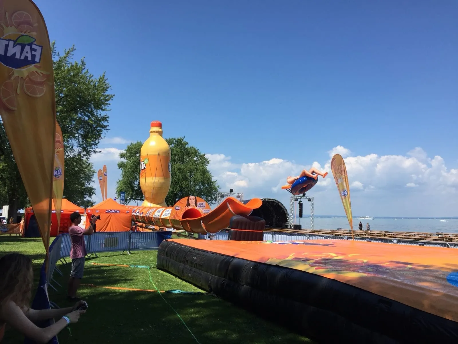 Orange Fanta water slide on a lakeshore, with a person flying out of the tube towards the air cushion