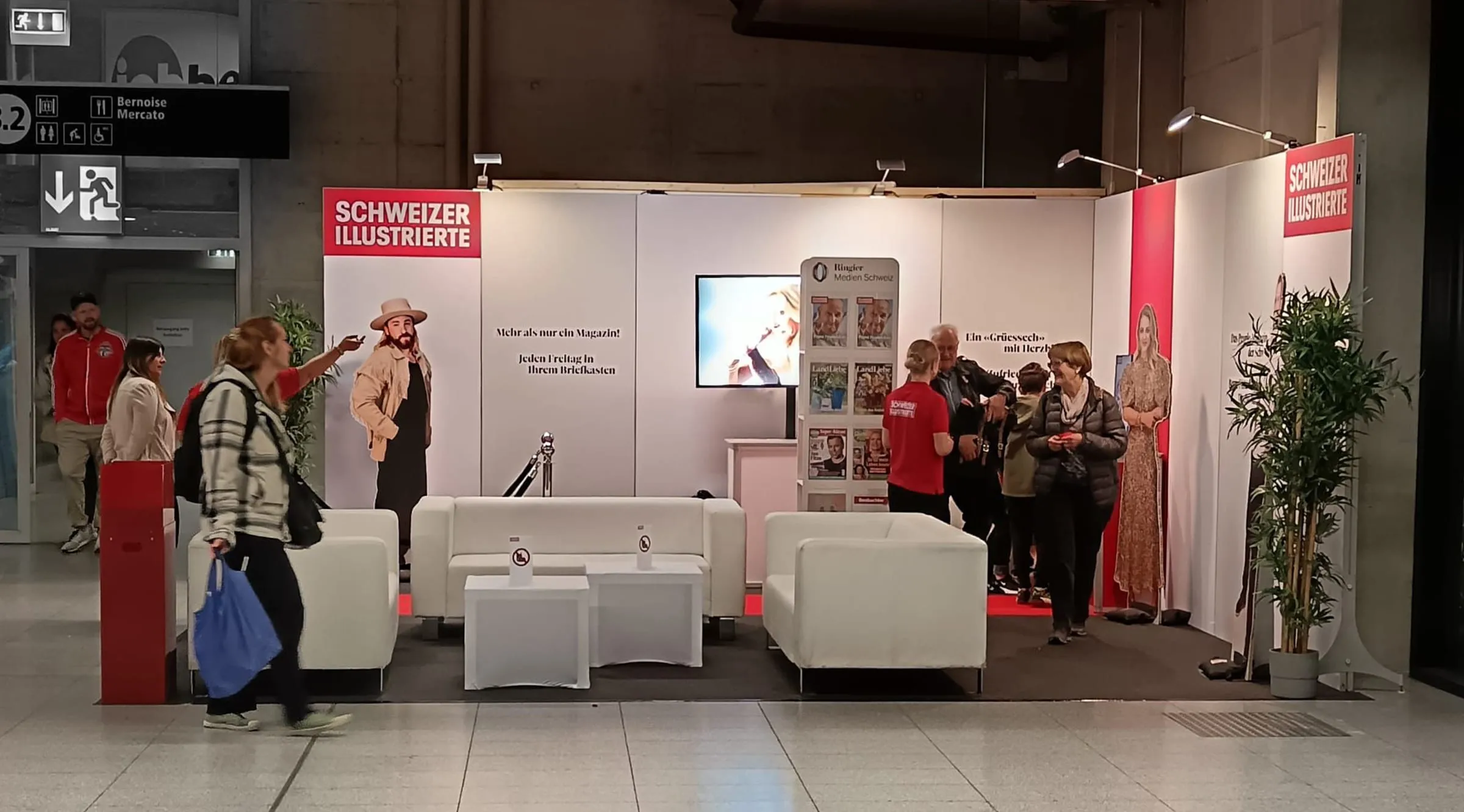 SI stand with white sofas, celebrity cutouts, a screen and magazine rack, with visitors walking past