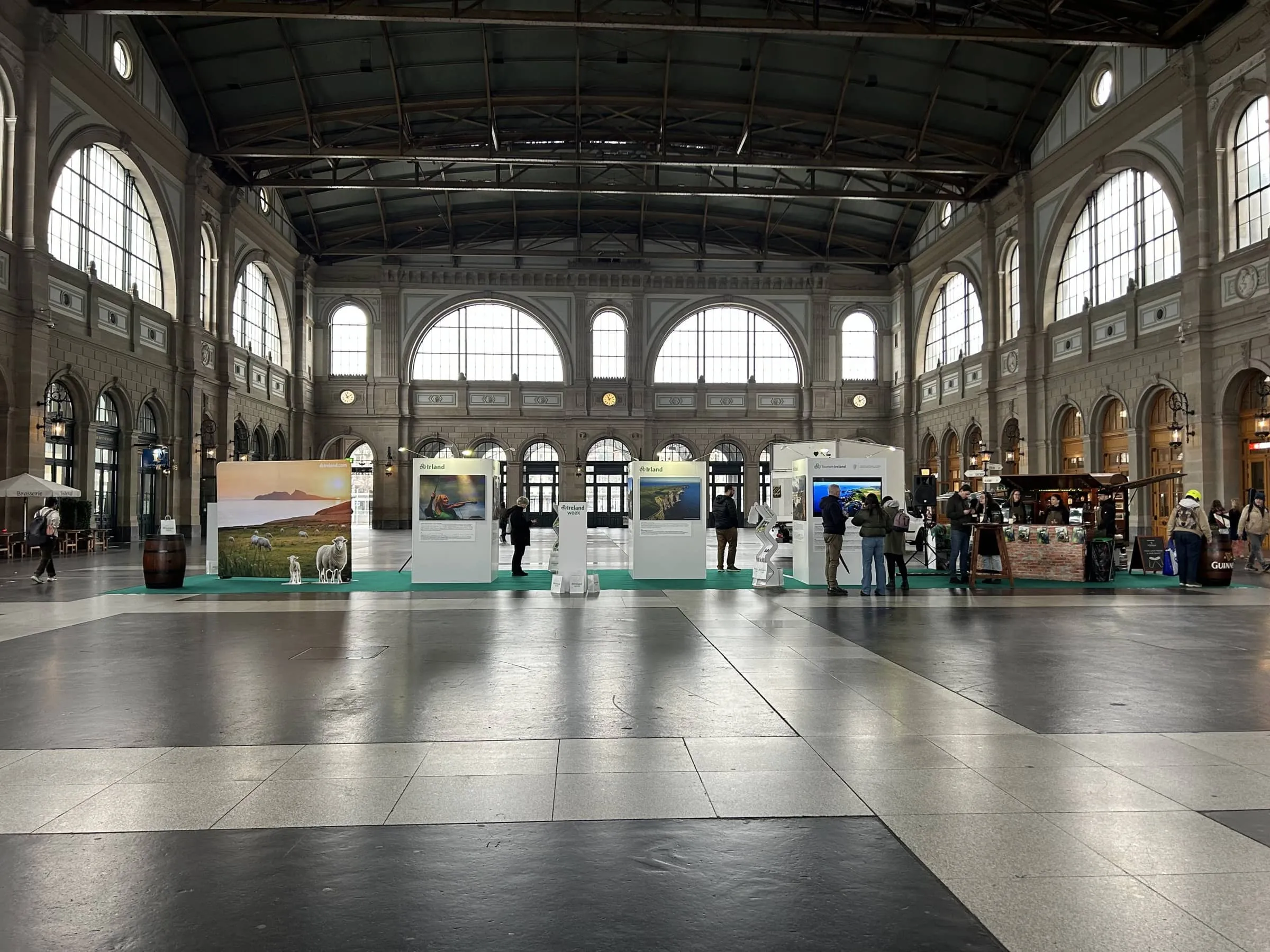 Photo exhibition on a green carpet in Zürich main station, with white display walls, cardboard sheep and an Irish pub
