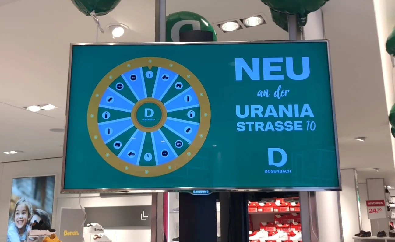 Screen showing a green-and-blue digital Glücksrad and the text «Neu an der Uraniastrasse 10» at Dosenbach